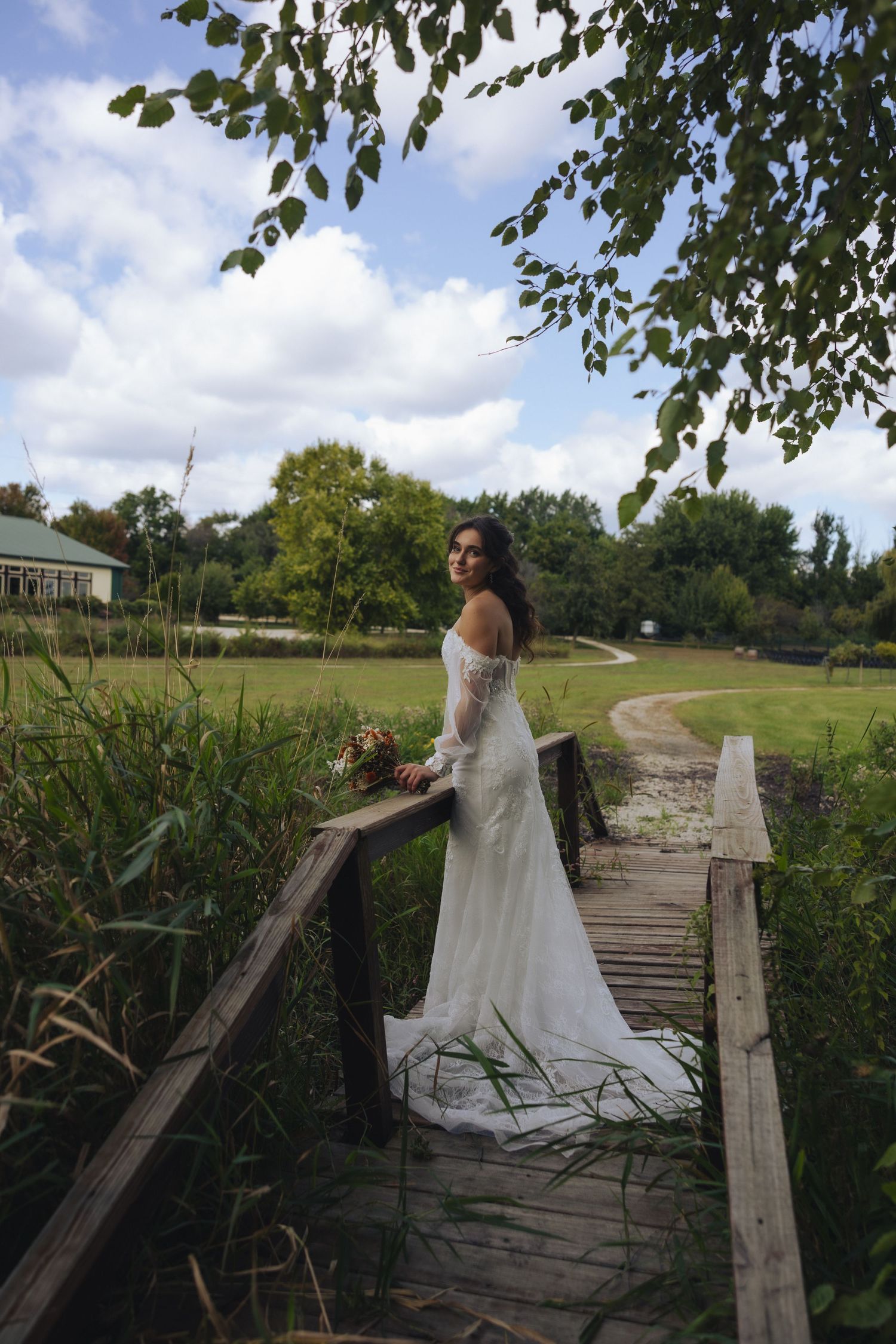 A person in a flowing white wedding dress poses on a rustic wooden bridge in a natural outdoor setting.