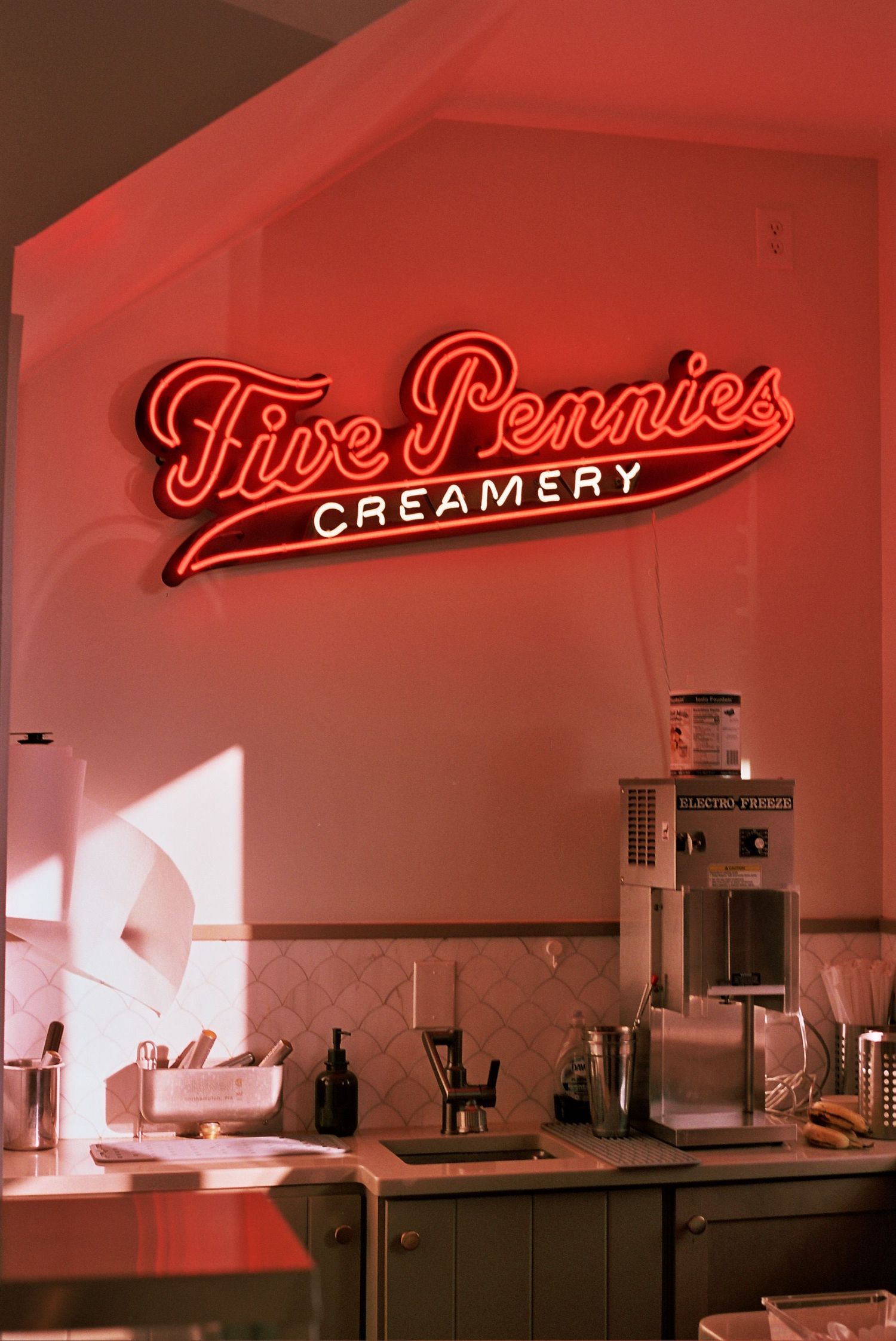 Retro neon 'Five Pennies Creamery' sign glows red above a counter with vintage equipment.