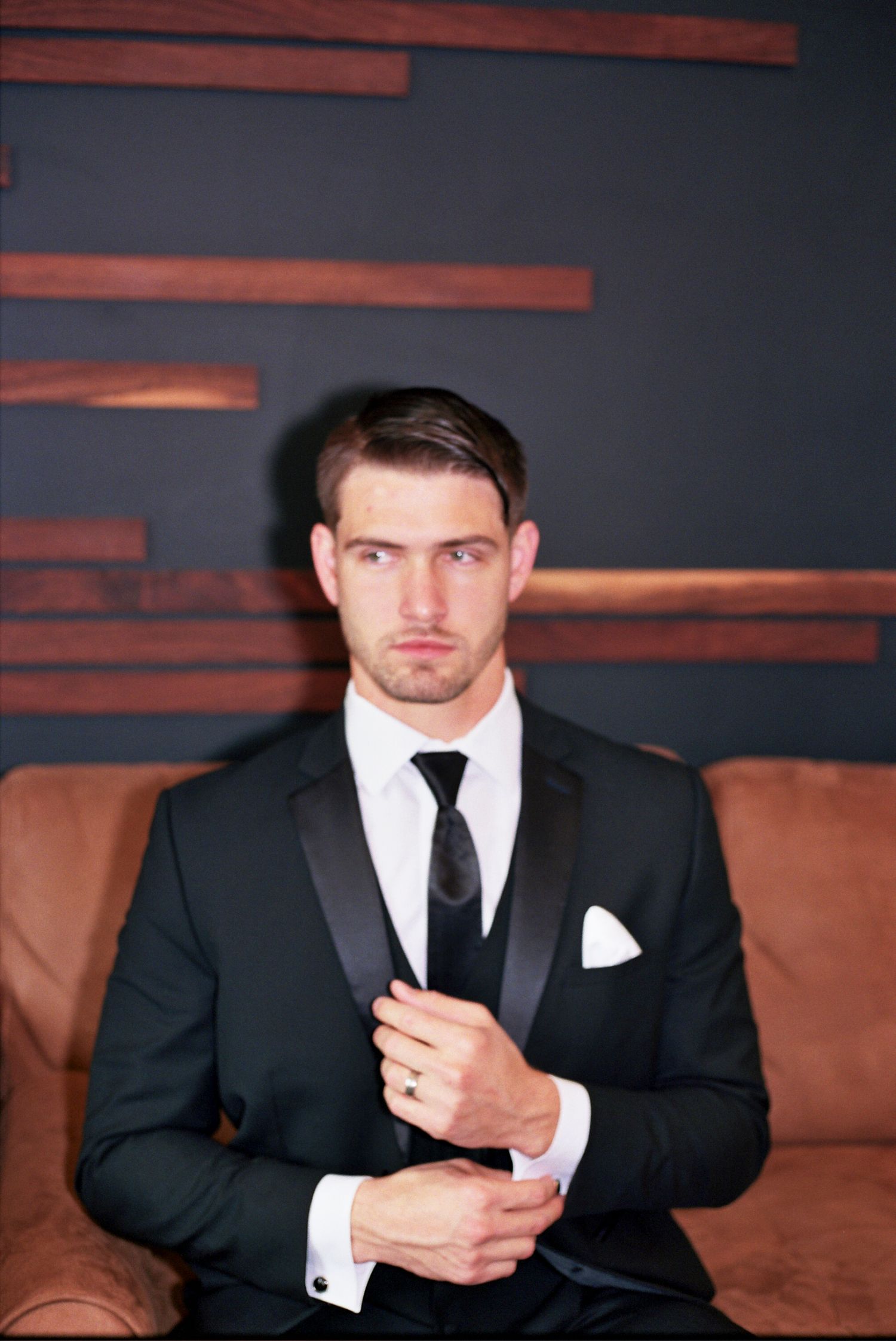 A person wearing a black suit and tie adjusts their cufflinks while seated in a dimly lit room.