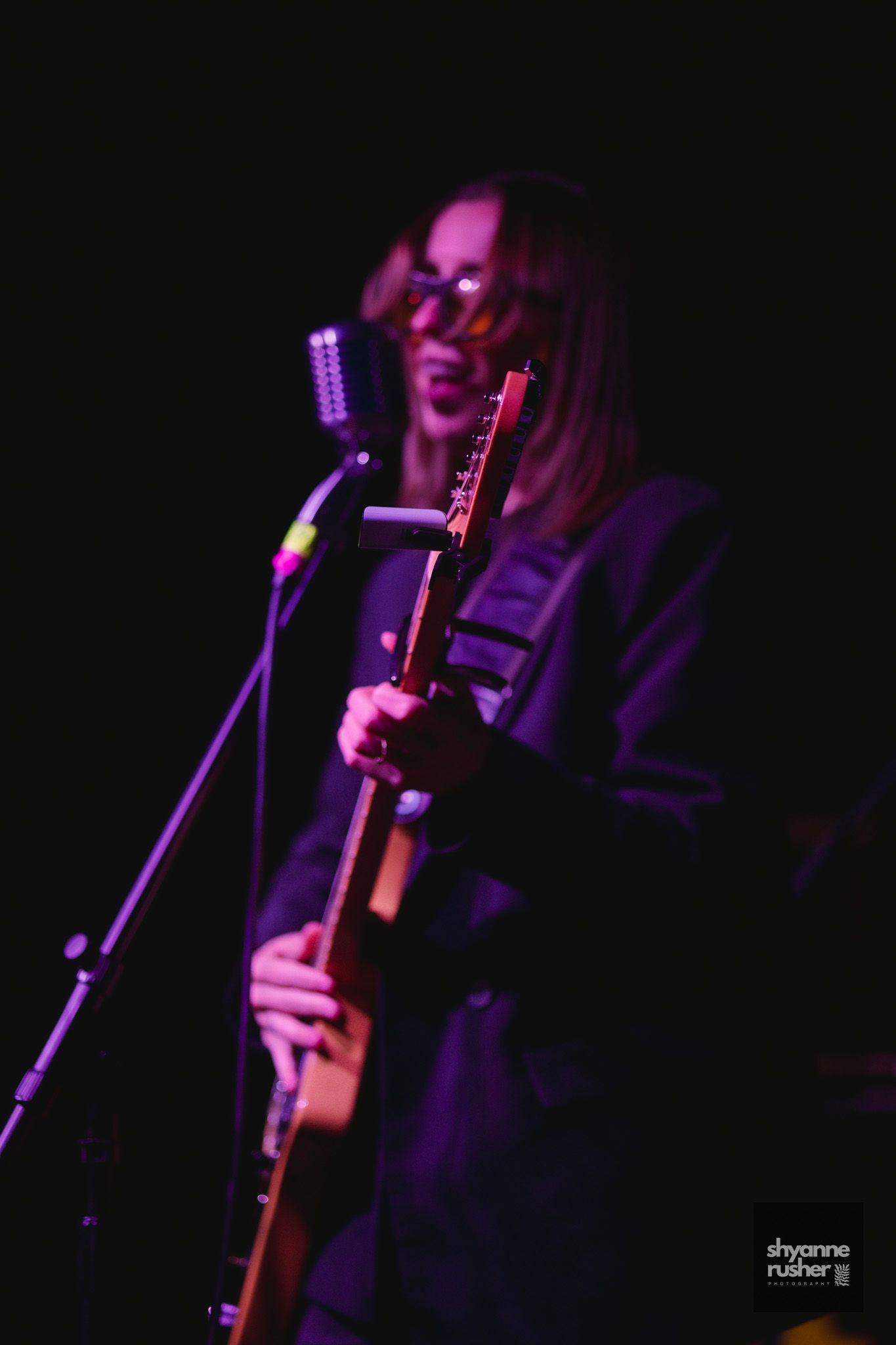 A musician performs on stage with a guitar under dramatic purple lighting at a concert.