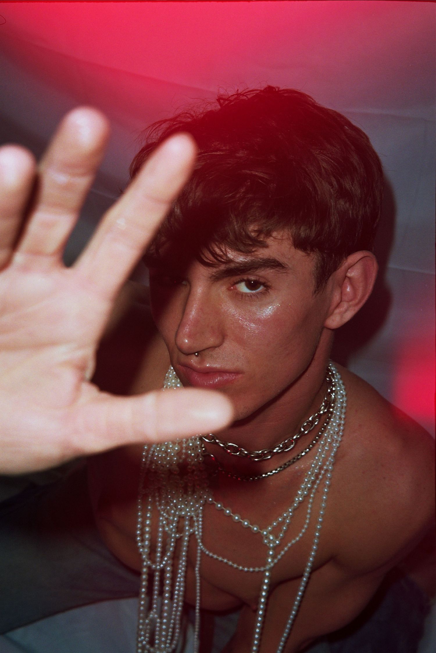 Young man with chain necklaces extends hand toward camera under dramatic red lighting.