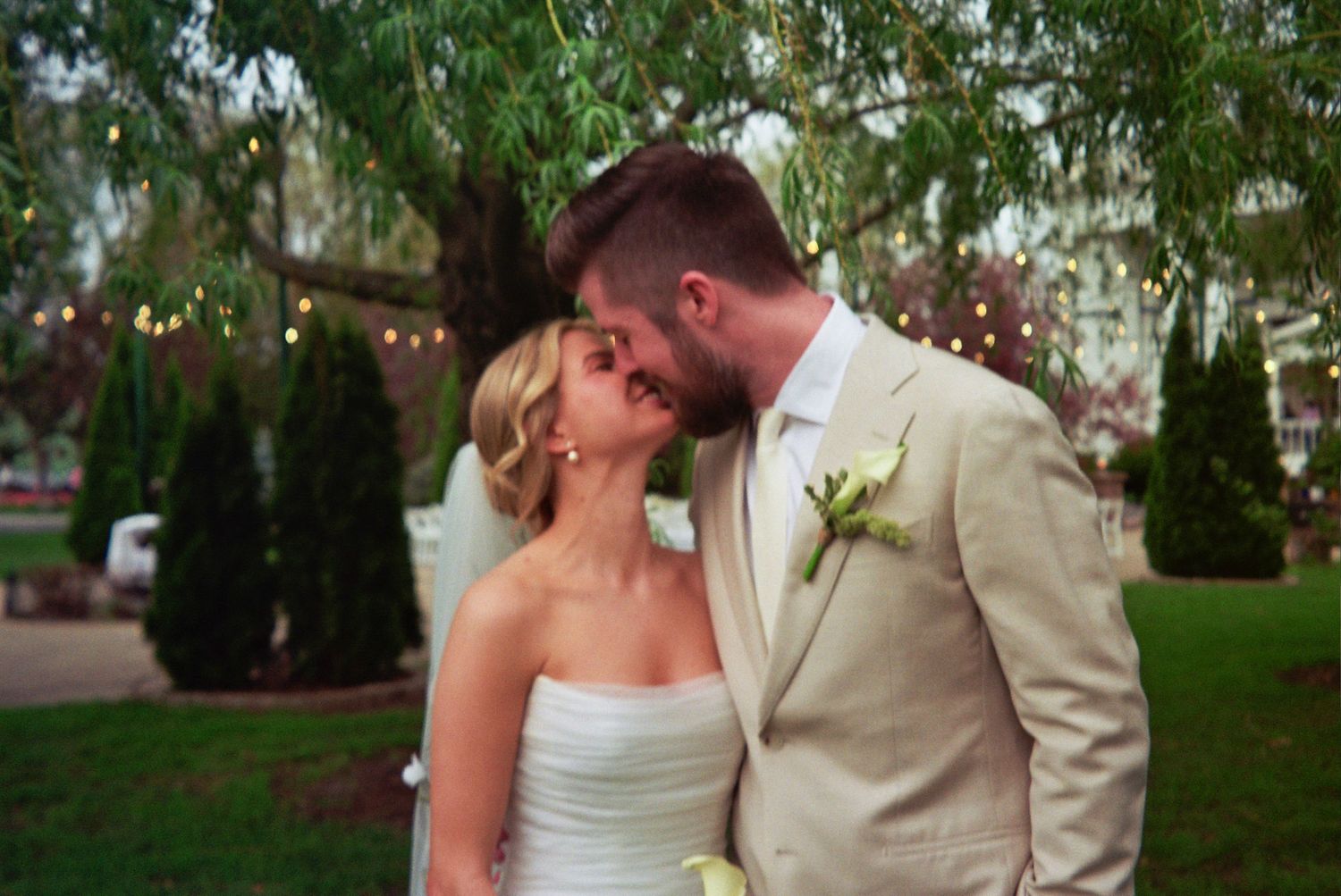 Bride and groom sharing a romantic kiss outdoors at a garden wedding under a willow tree with string lights.