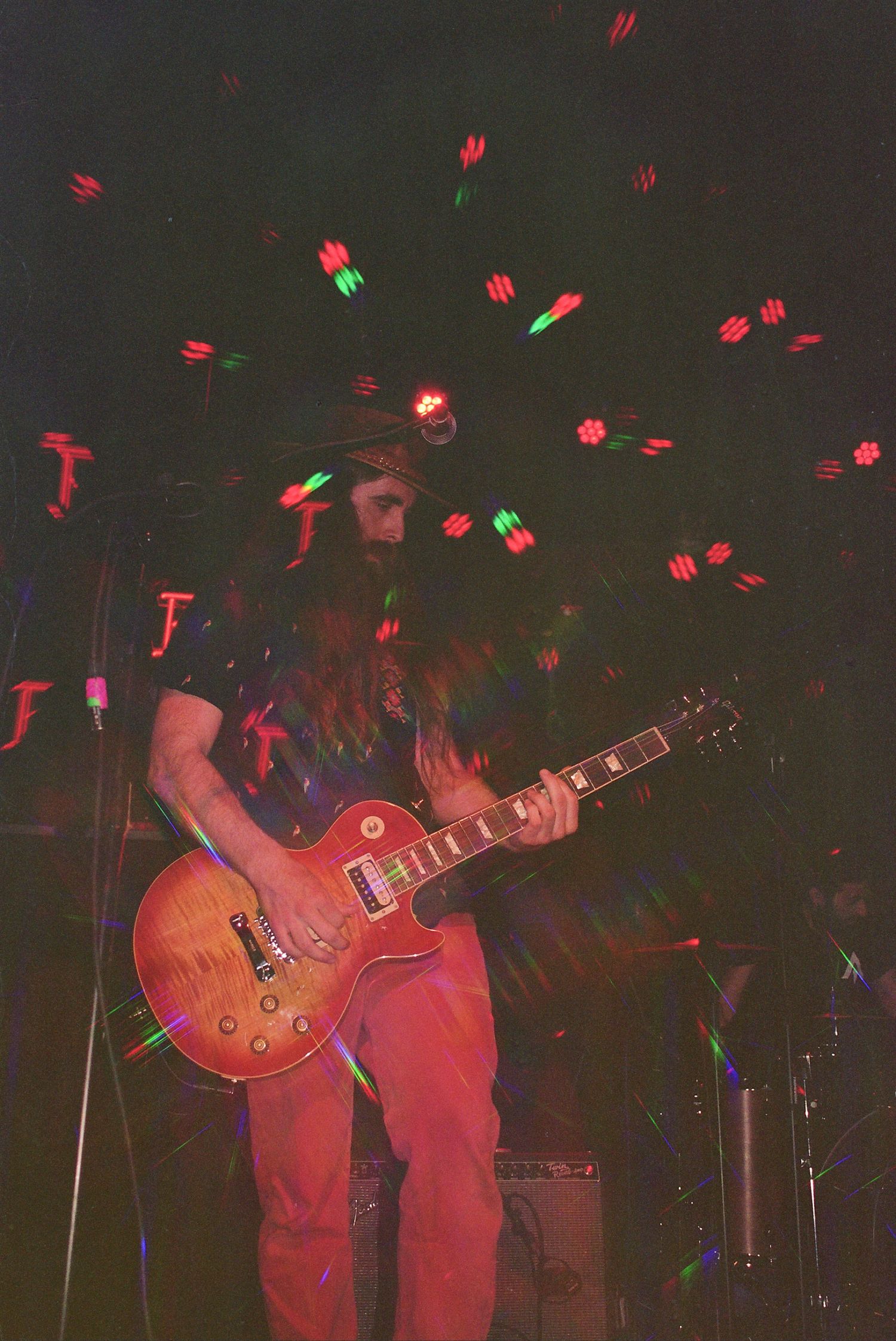 Bearded guitarist performing on stage with colorful bokeh lights, playing a sunburst Les Paul electric guitar under dramatic lighting.