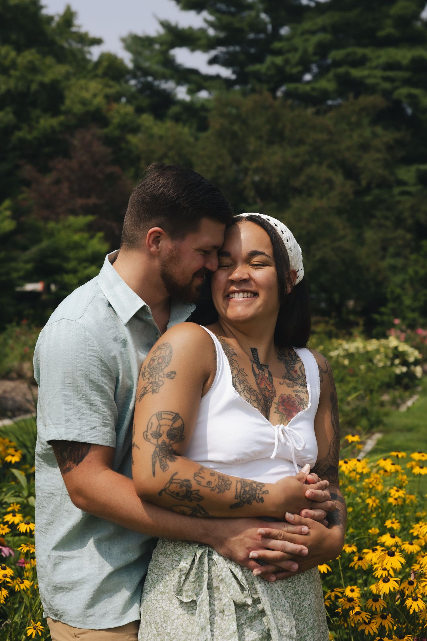 A couple embraces in a flower garden, the tattooed woman smiling joyfully as her partner whispers in her ear.