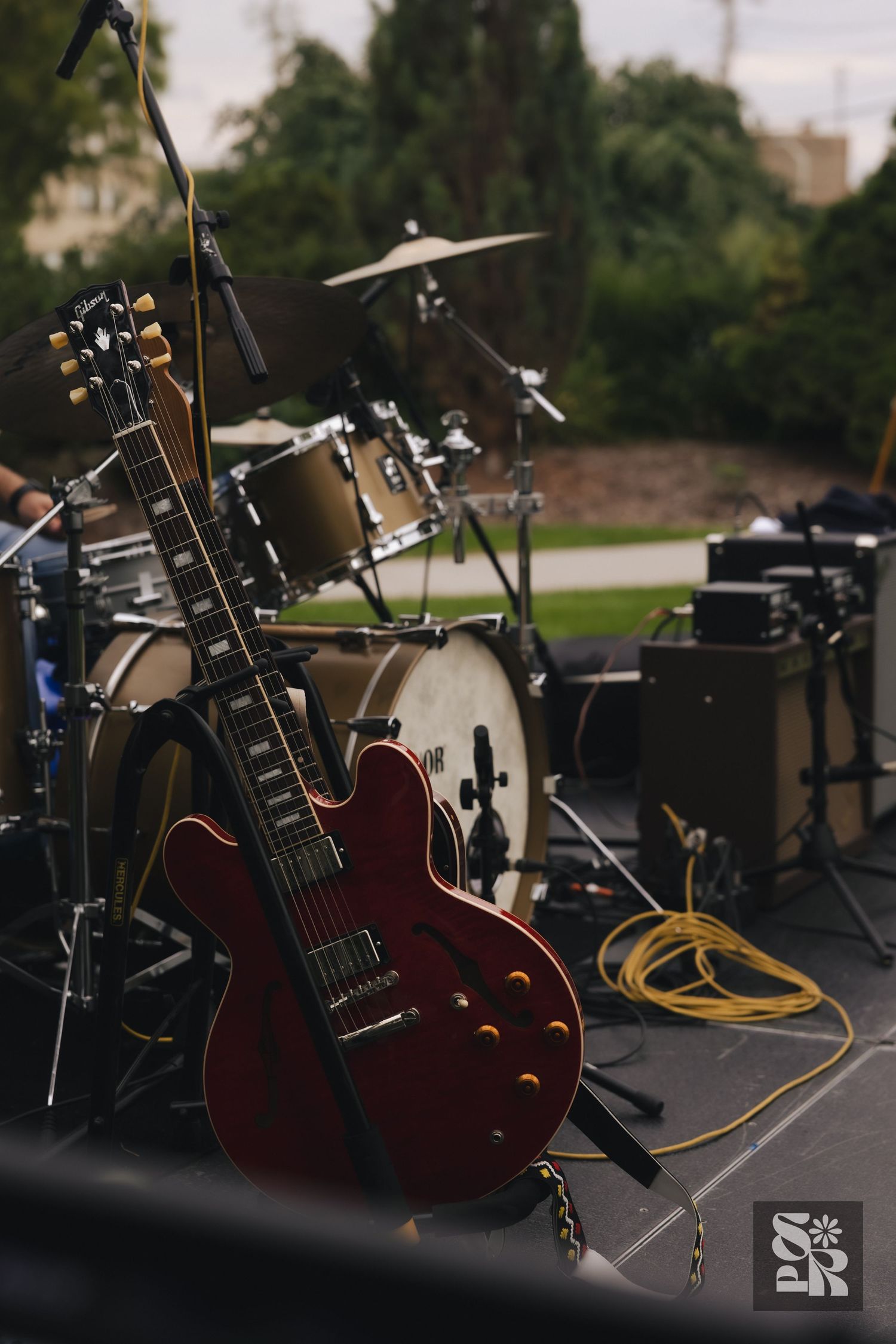 Red semi-hollow electric guitar on stage with drum kit and green trees in background at outdoor concert.