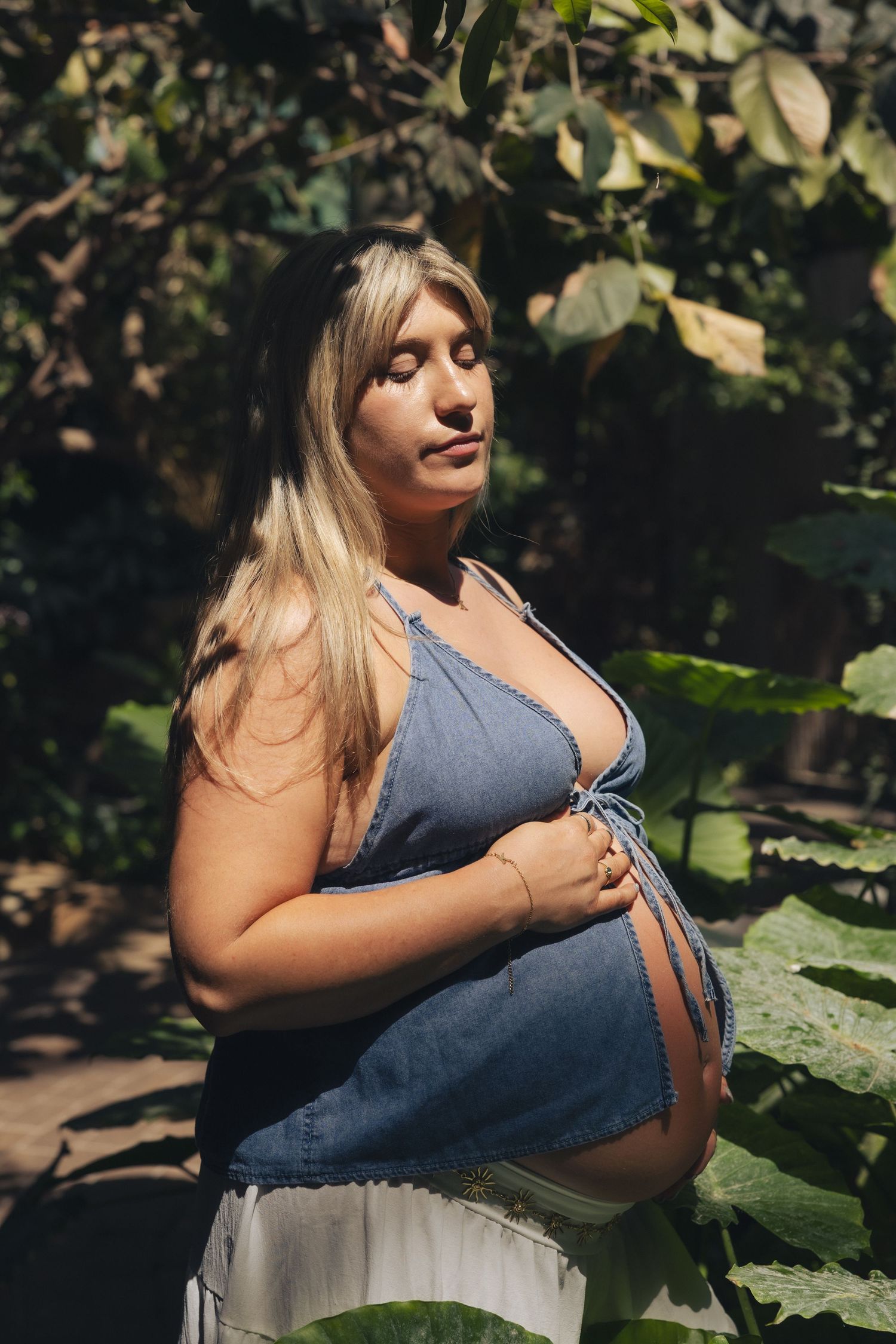 Pregnant woman with blonde hair in blue denim top holding baby bump, standing outdoors surrounded by lush green foliage.