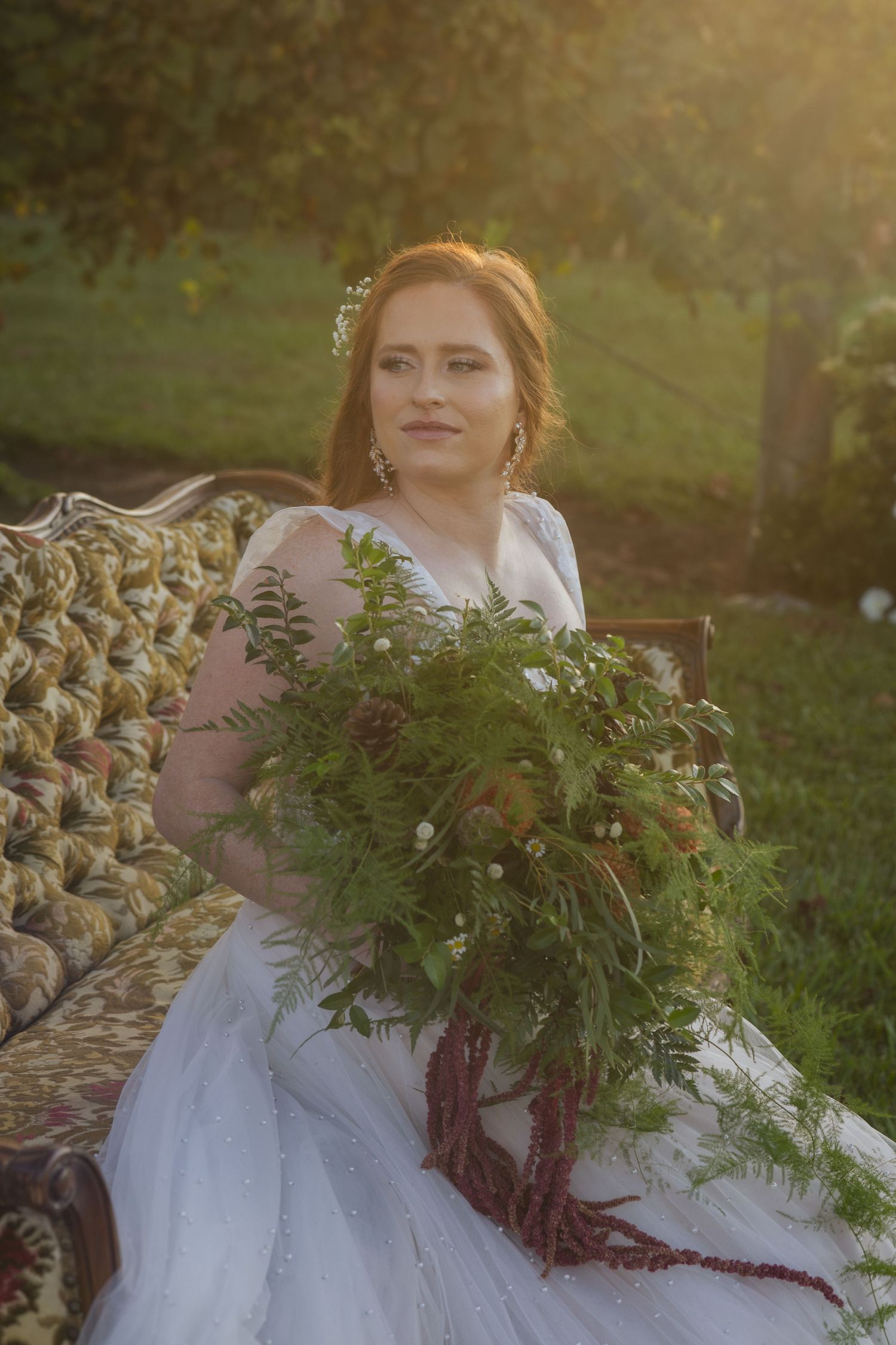 A bride in a white dress sits on a woven chair holding a lush greenery bouquet during golden hour lighting outdoors.