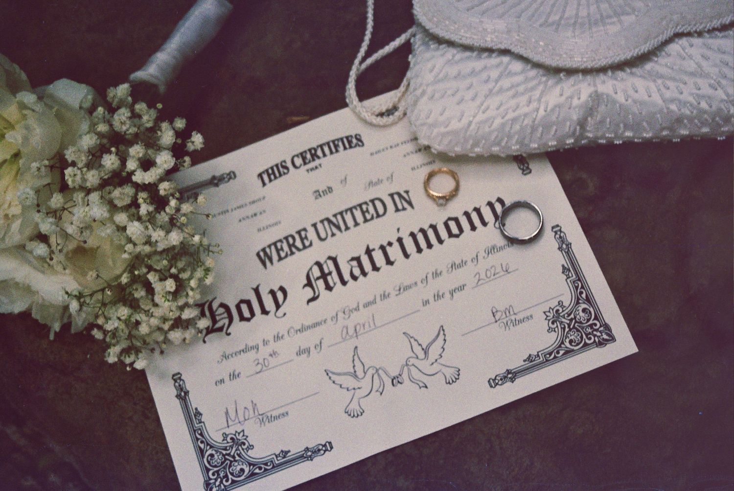 Holy matrimony marriage certificate with wedding rings, bridal bouquet, and lace veil on a dark surface.