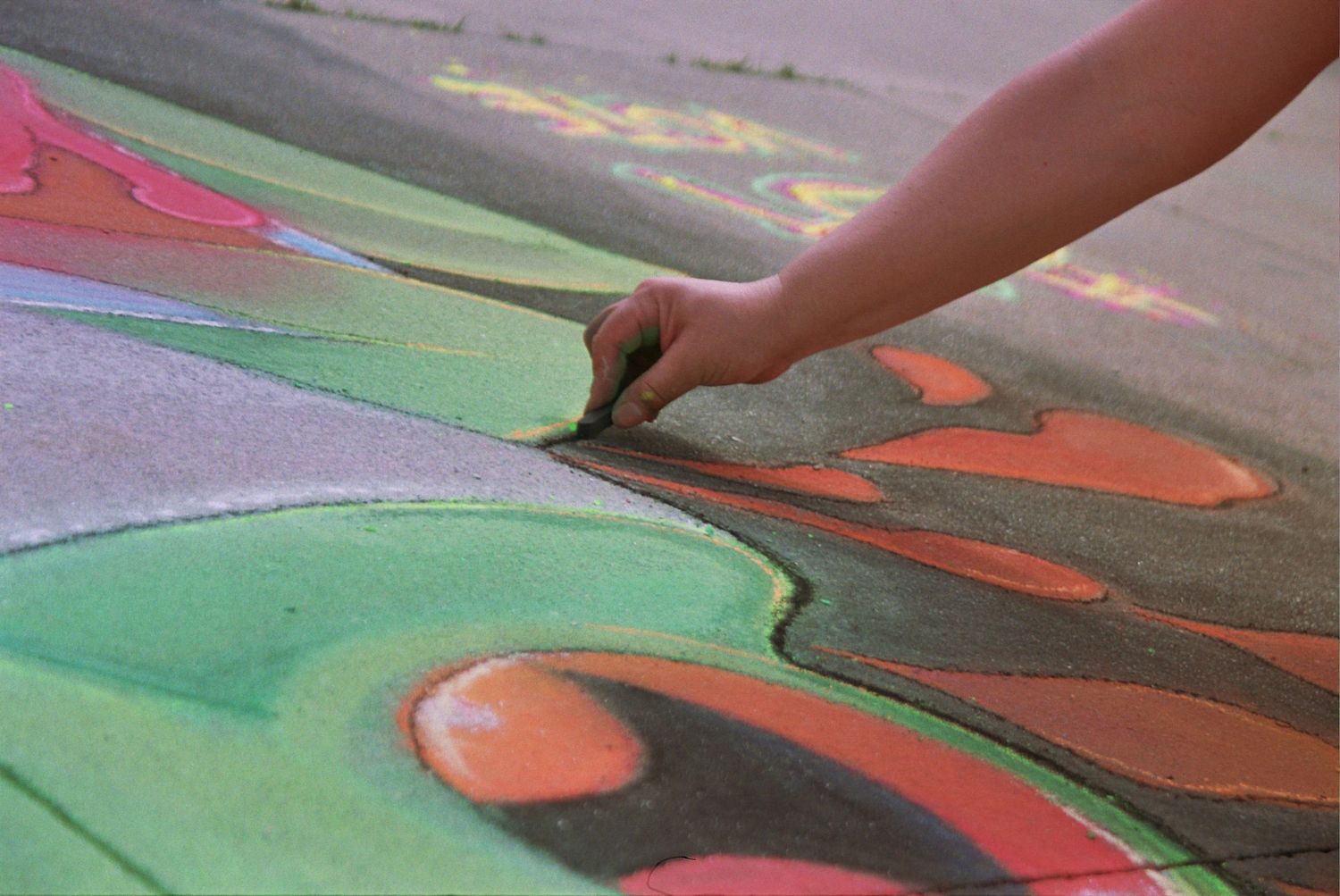 Child's hand drawing colorful chalk art on pavement, featuring green, red, and orange abstract shapes.