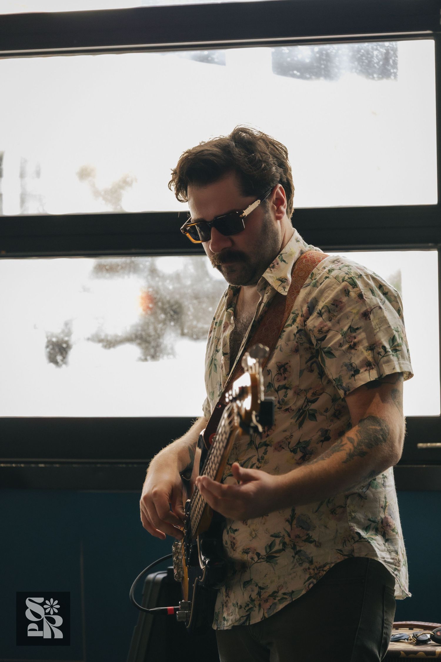 Tattooed musician in floral shirt and sunglasses playing electric guitar indoors near a window.