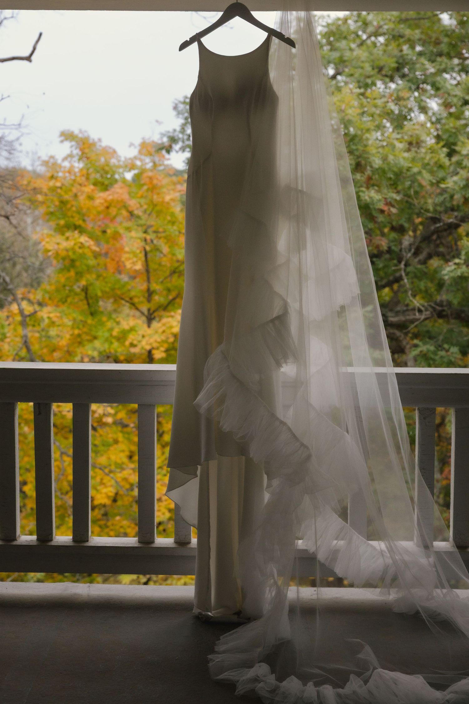 A flowing white wedding dress hangs elegantly on a balcony railing with autumn foliage visible in the background.