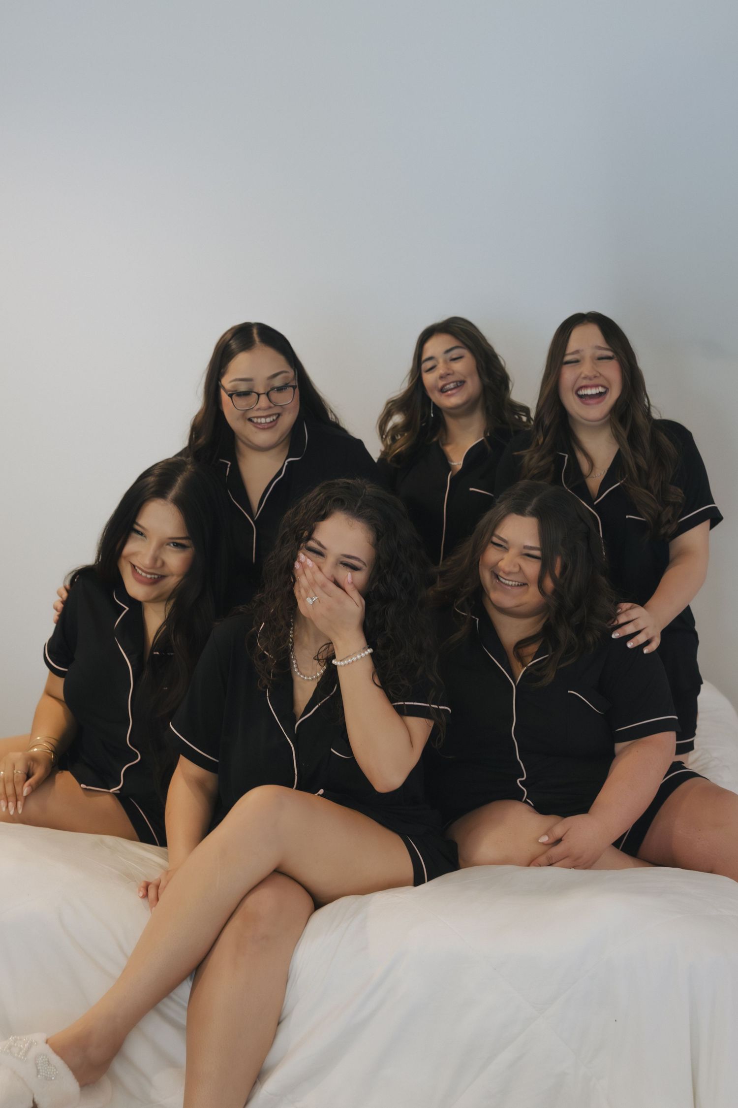 A group of friends in matching black pajamas pose together on white bedding during a sleepover photoshoot.