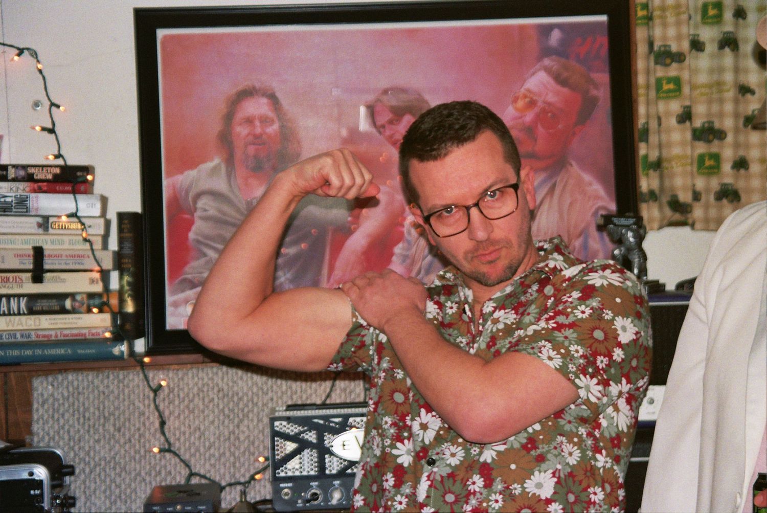 Man in floral shirt flexing bicep indoors with colorful painting of figures hanging on wall behind him.