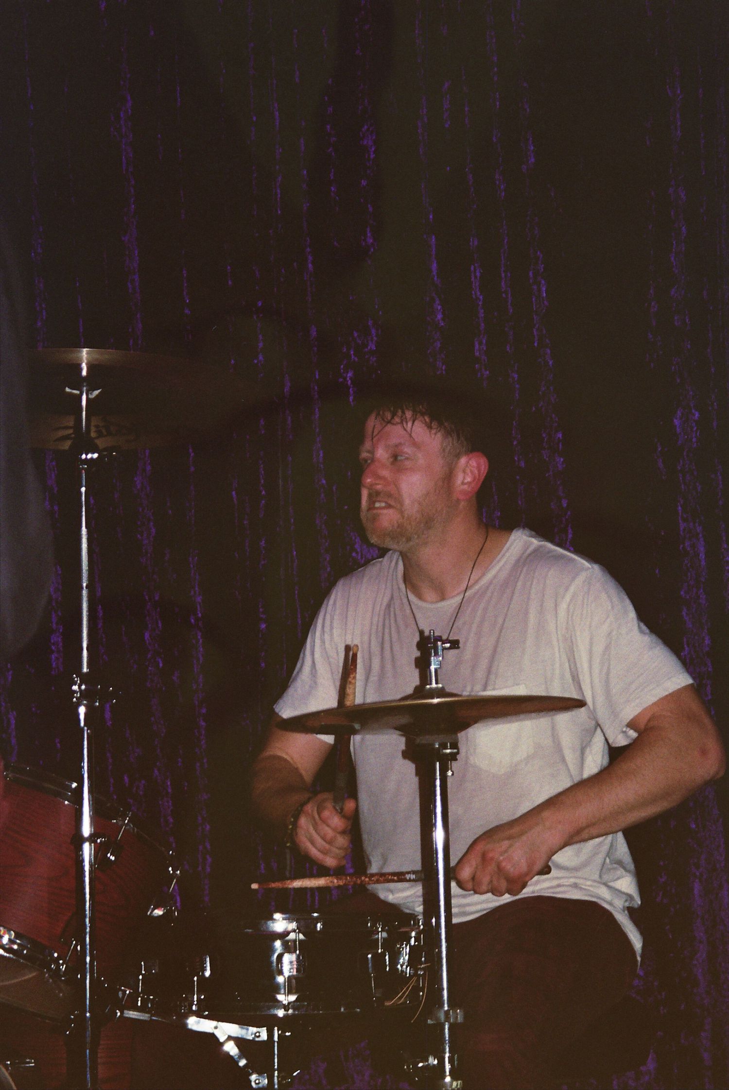 A male drummer performs on stage in a white t-shirt, playing cymbals with drumsticks under dim purple lighting.