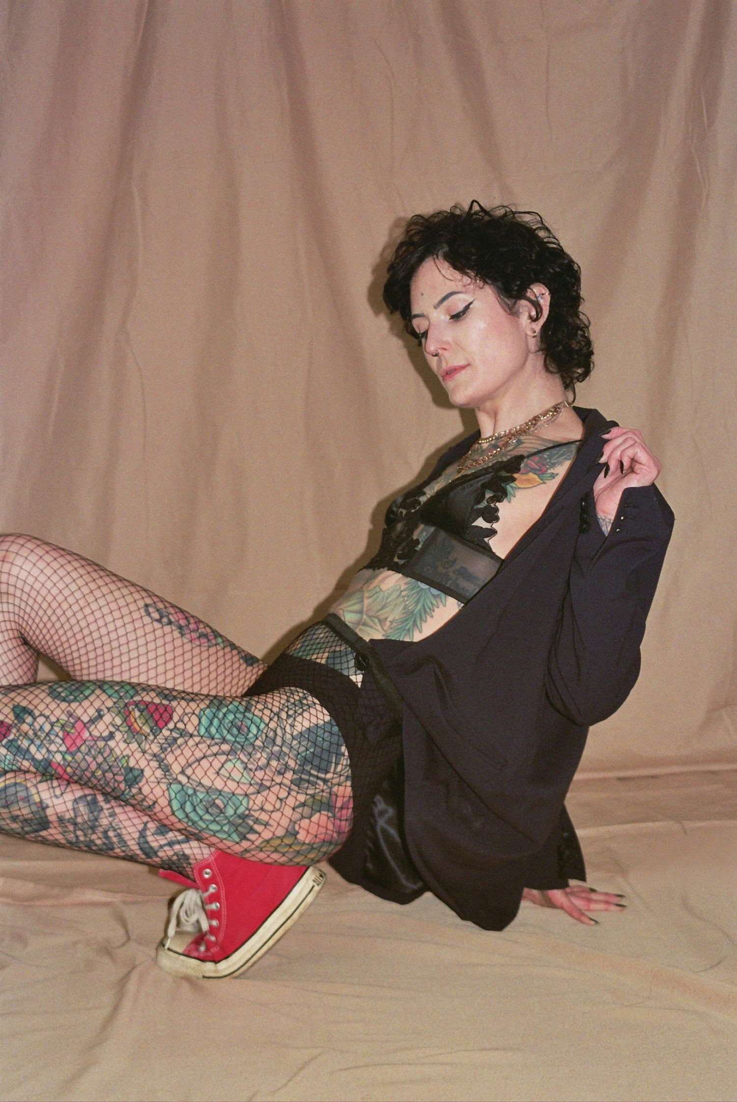 Tattooed woman in fishnet tights, black lingerie, blazer, and red Converse sneakers poses against beige backdrop.