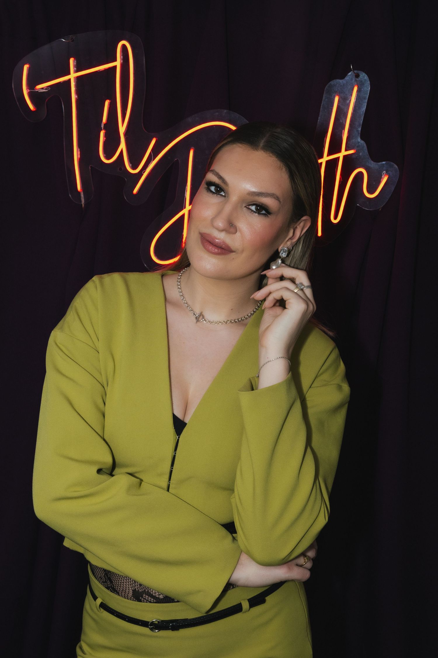 Woman in yellow-green blazer poses before neon 'Til Death' sign, wearing silver jewelry and elegant makeup.