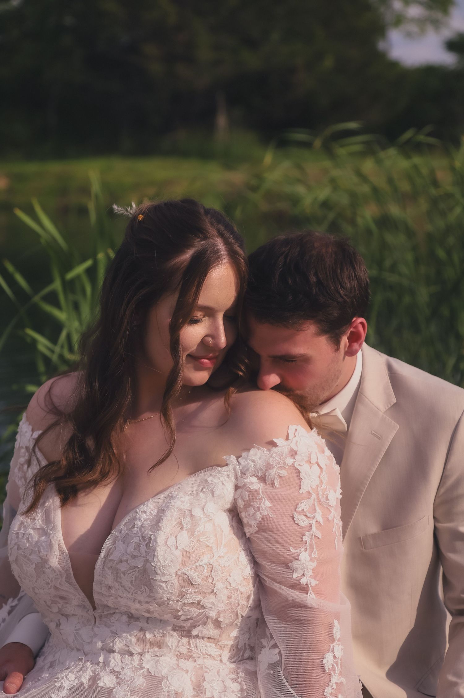 A tender intimate moment between a couple in wedding attire, with lace details on the dress and a light beige suit.