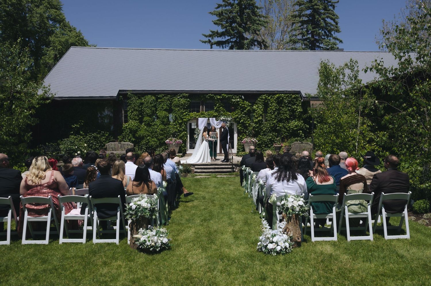 Outdoor wedding ceremony on green lawn with guests seated in white chairs facing couple at floral arch beside ivy-covered building.