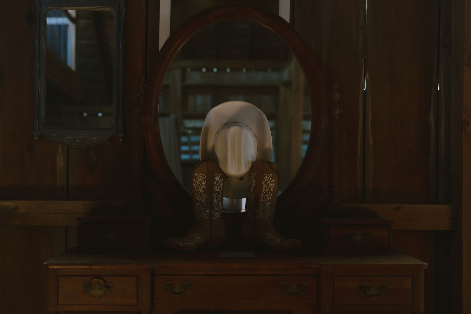 A dimly lit wooden dresser with an oval mirror reflecting warm light in a dark moody interior.