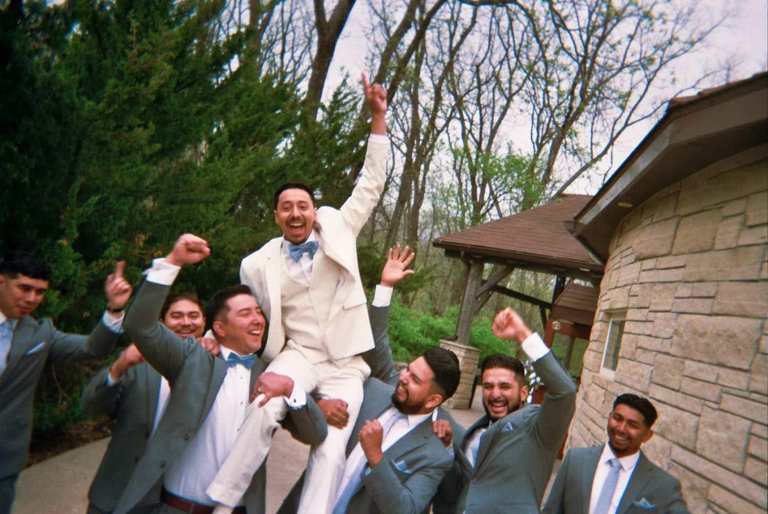 A group of groomsmen in gray suits lift and celebrate with excitement outside a stone building.