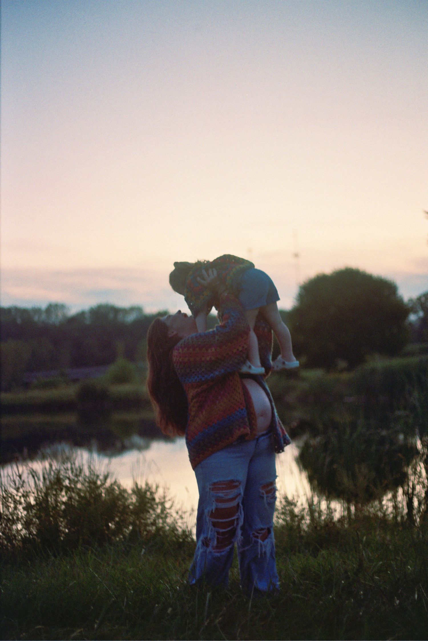 A silhouetted figure holds a small child at sunset by a peaceful lake, creating a serene family moment.