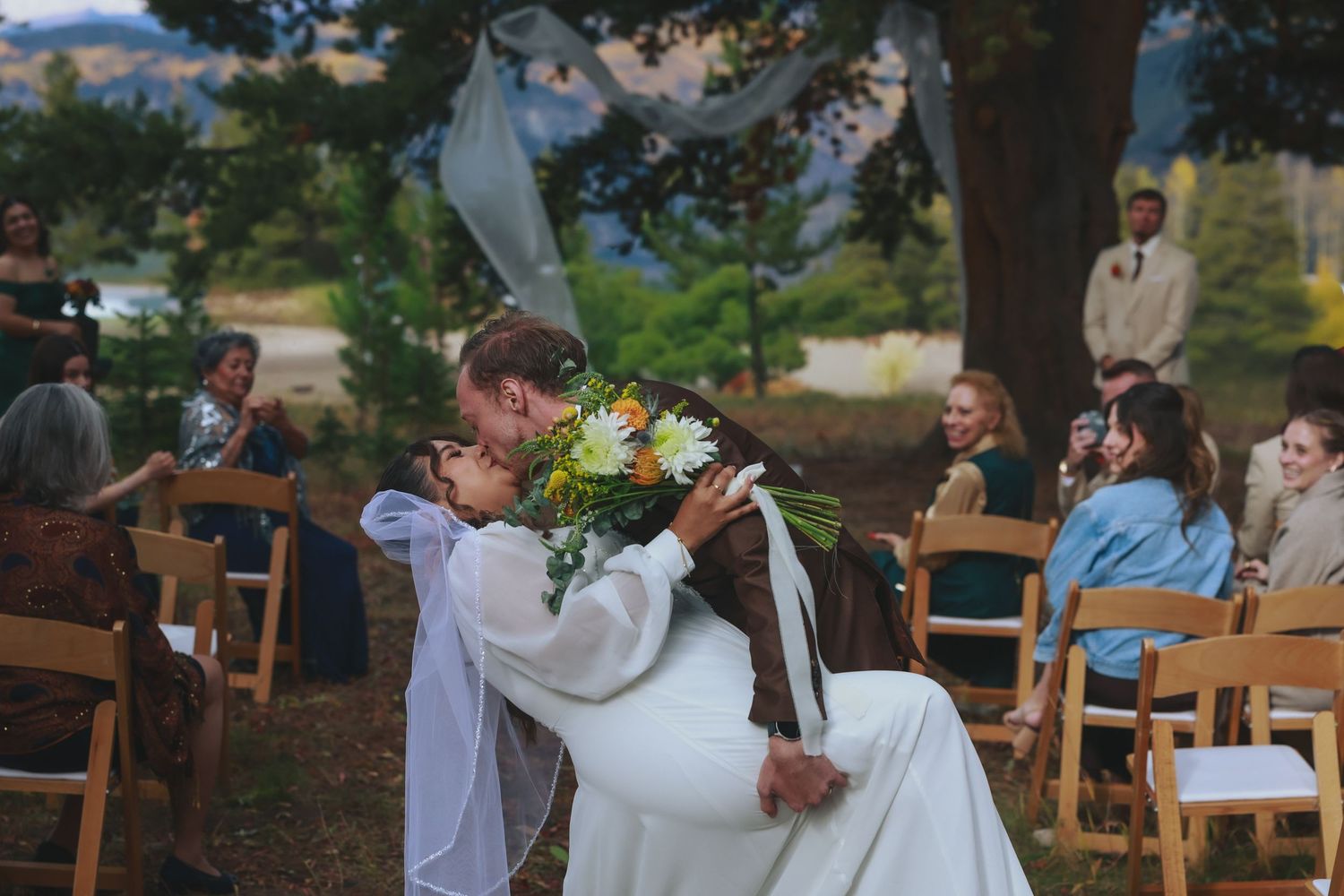 A couple shares a tender moment during their outdoor wedding ceremony surrounded by wooden chairs.