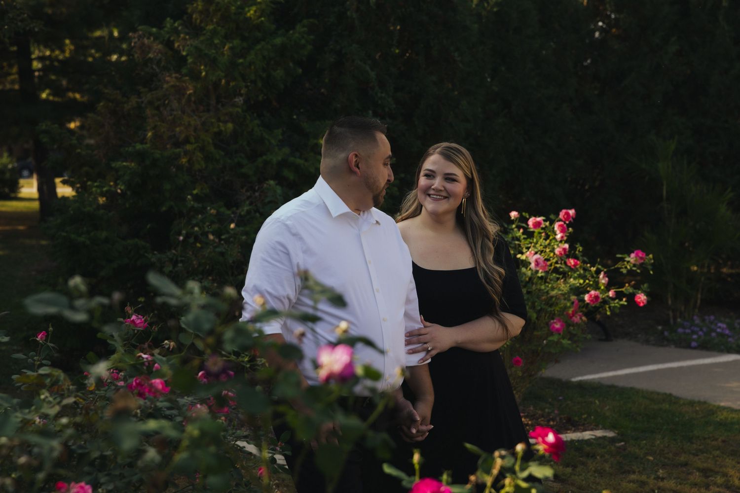 Couple smiling together in a garden surrounded by pink roses, man in white shirt and woman in black dress.