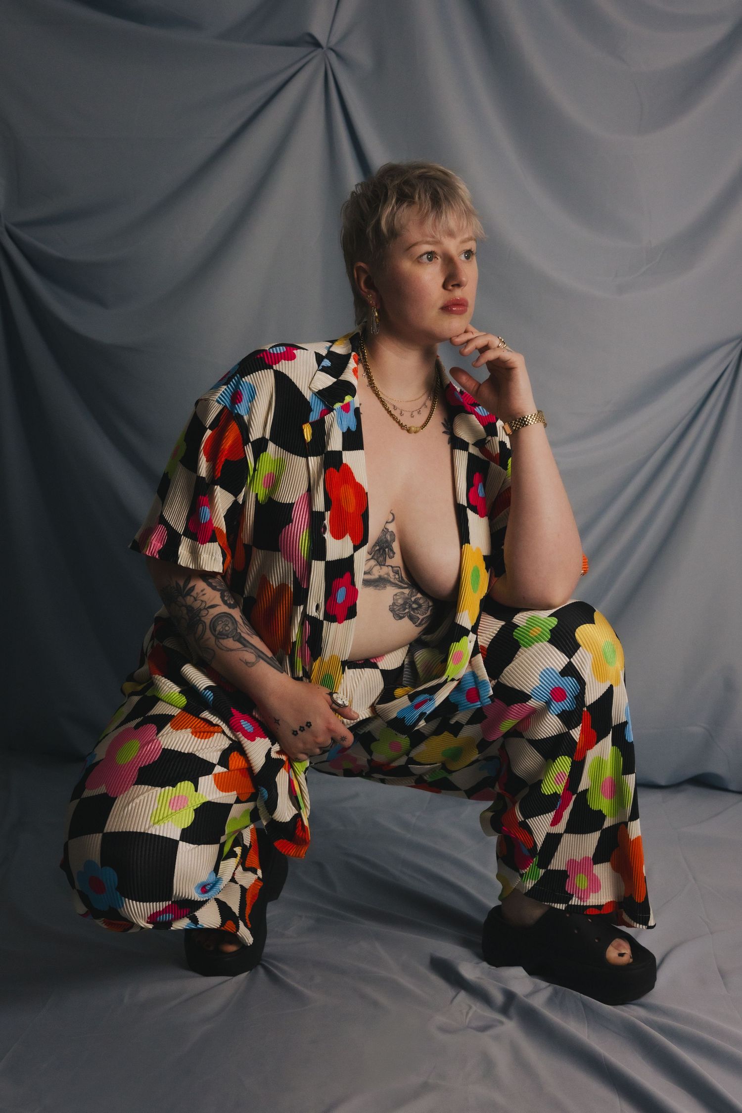 Person posing in colorful checkered floral wide-leg pantsuit with open shirt, seated against gray draped backdrop.