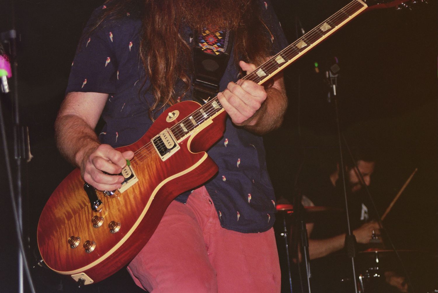 Guitarist with long hair plays a sunburst Les Paul electric guitar on stage under colorful lights.