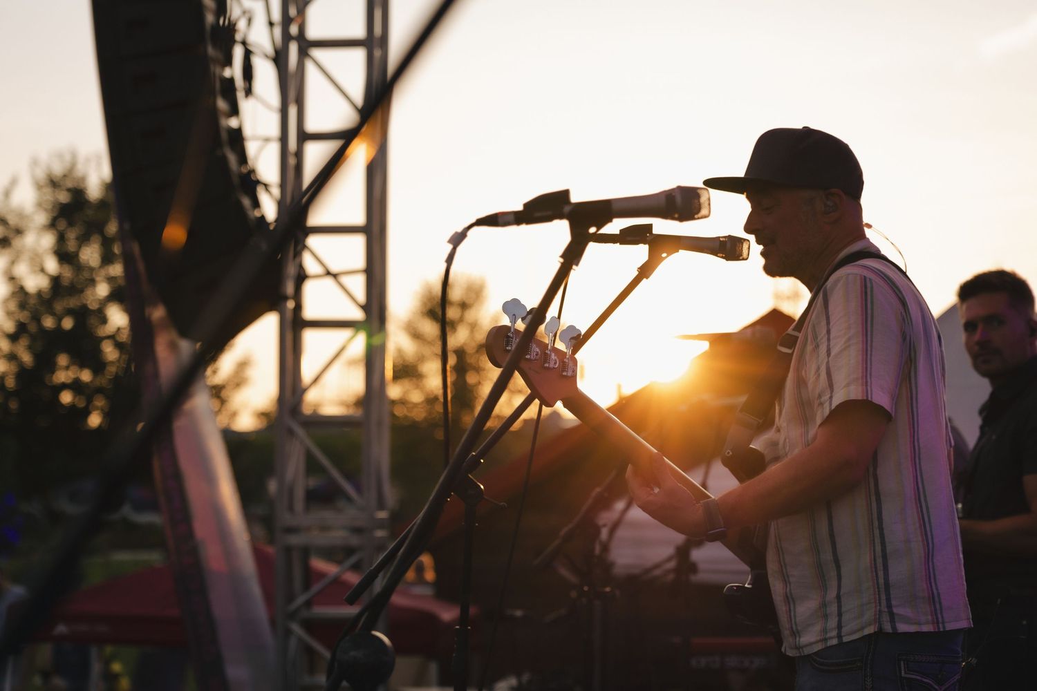Musician performing on outdoor stage at sunset with microphone and guitar silhouetted against golden sky.