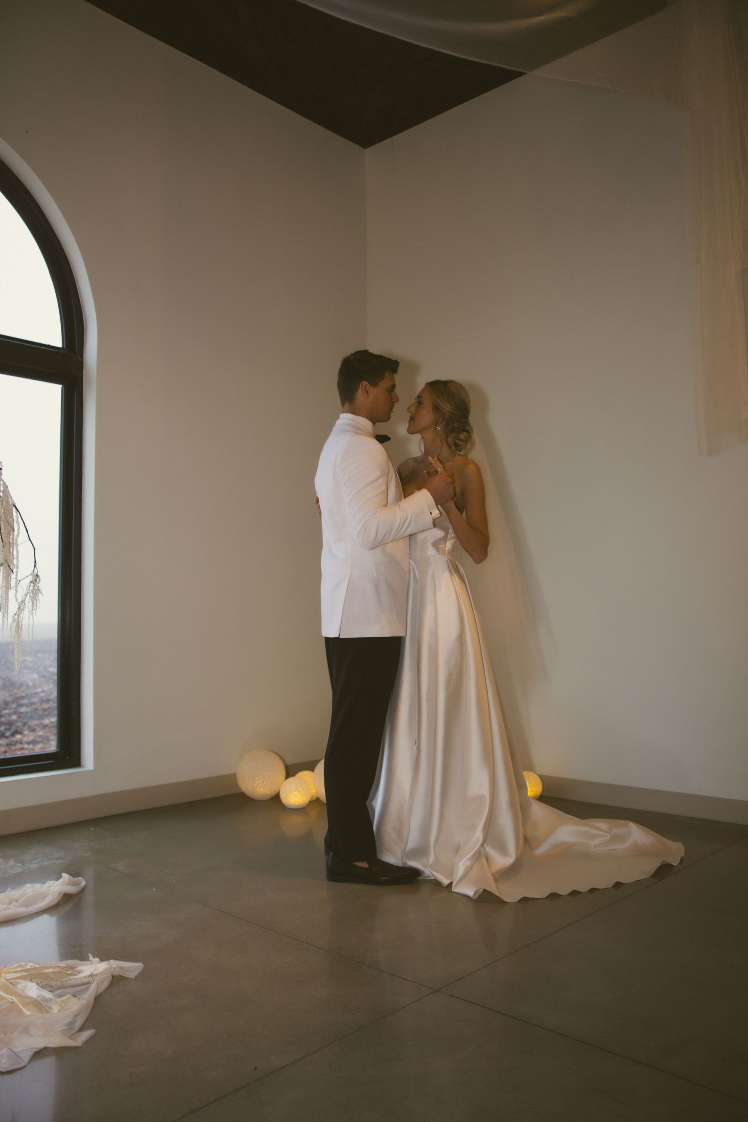 Romantic wedding couple shares an intimate moment in a minimalist room with arched windows and candlelight.