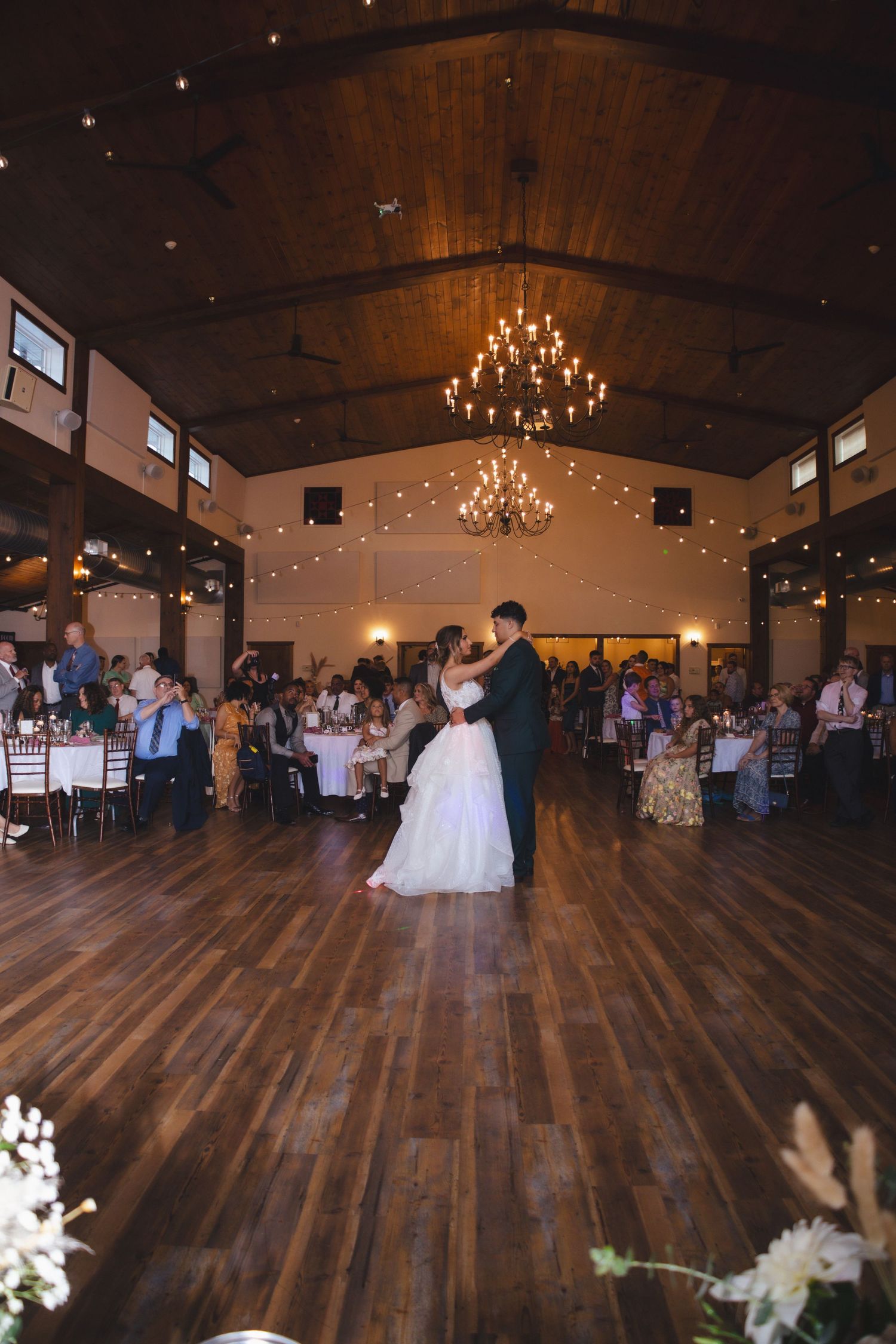First dance at wedding reception in rustic venue with wooden floors and chandeliers.