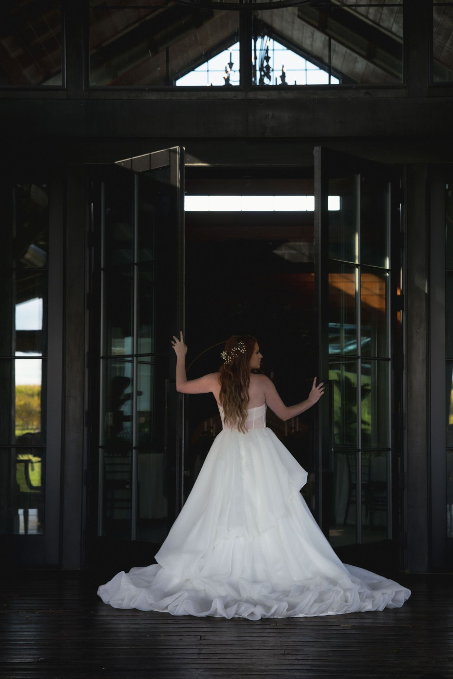 A bride in a flowing white gown stands silhouetted against glass doors showing her dramatic backless dress design.