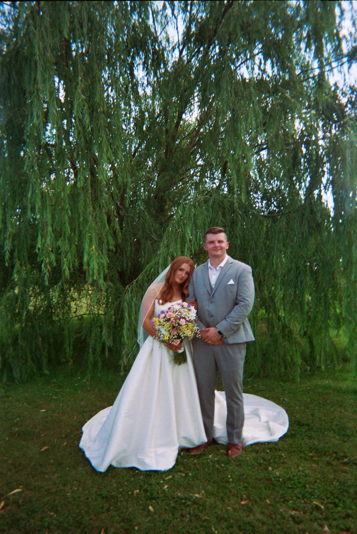 A happy couple poses under a beautiful weeping willow tree on a lovely outdoor wedding day.