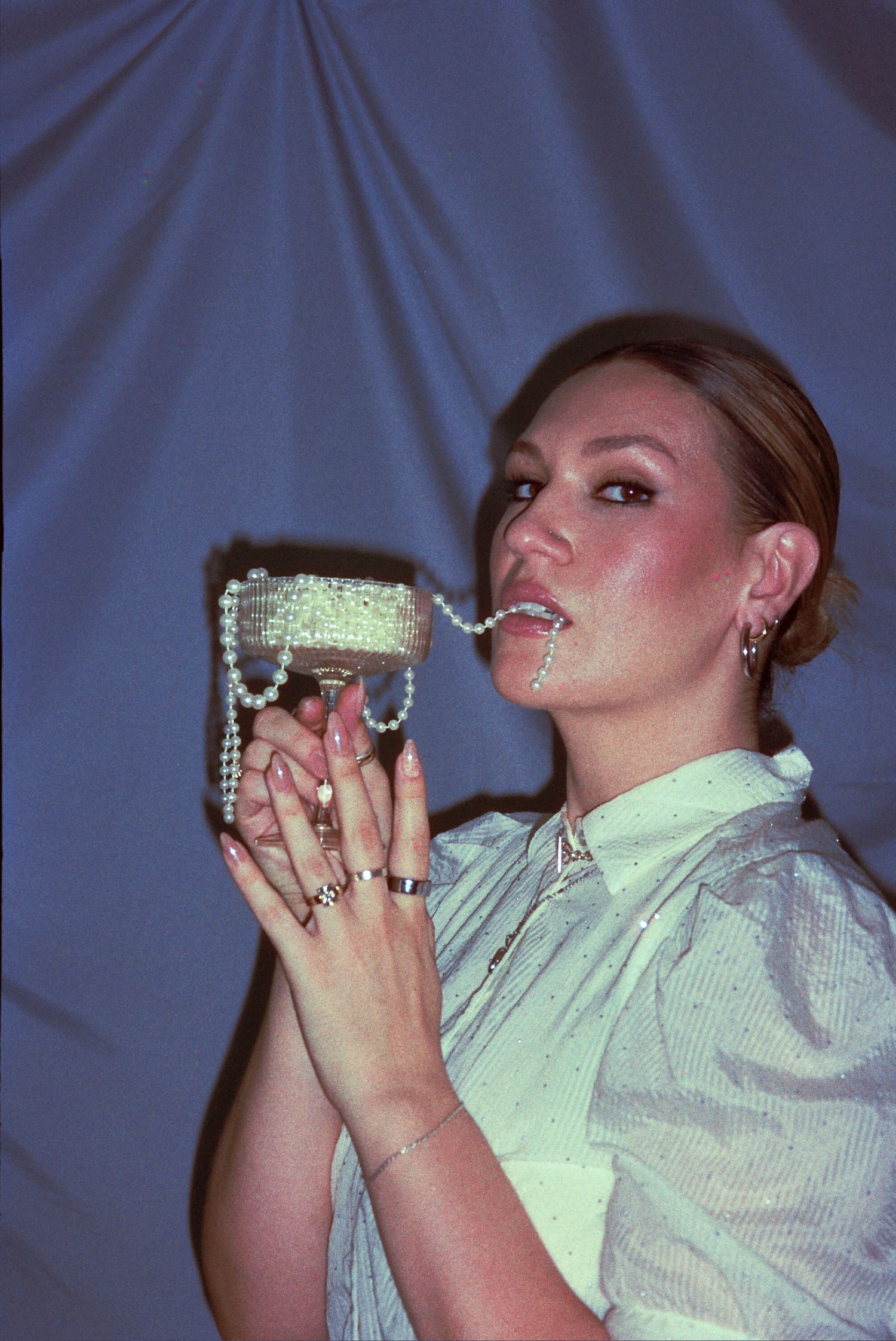 Woman posing with a crystal-embellished handbag, pearl chain in mouth, wearing rings and earrings against blue backdrop.