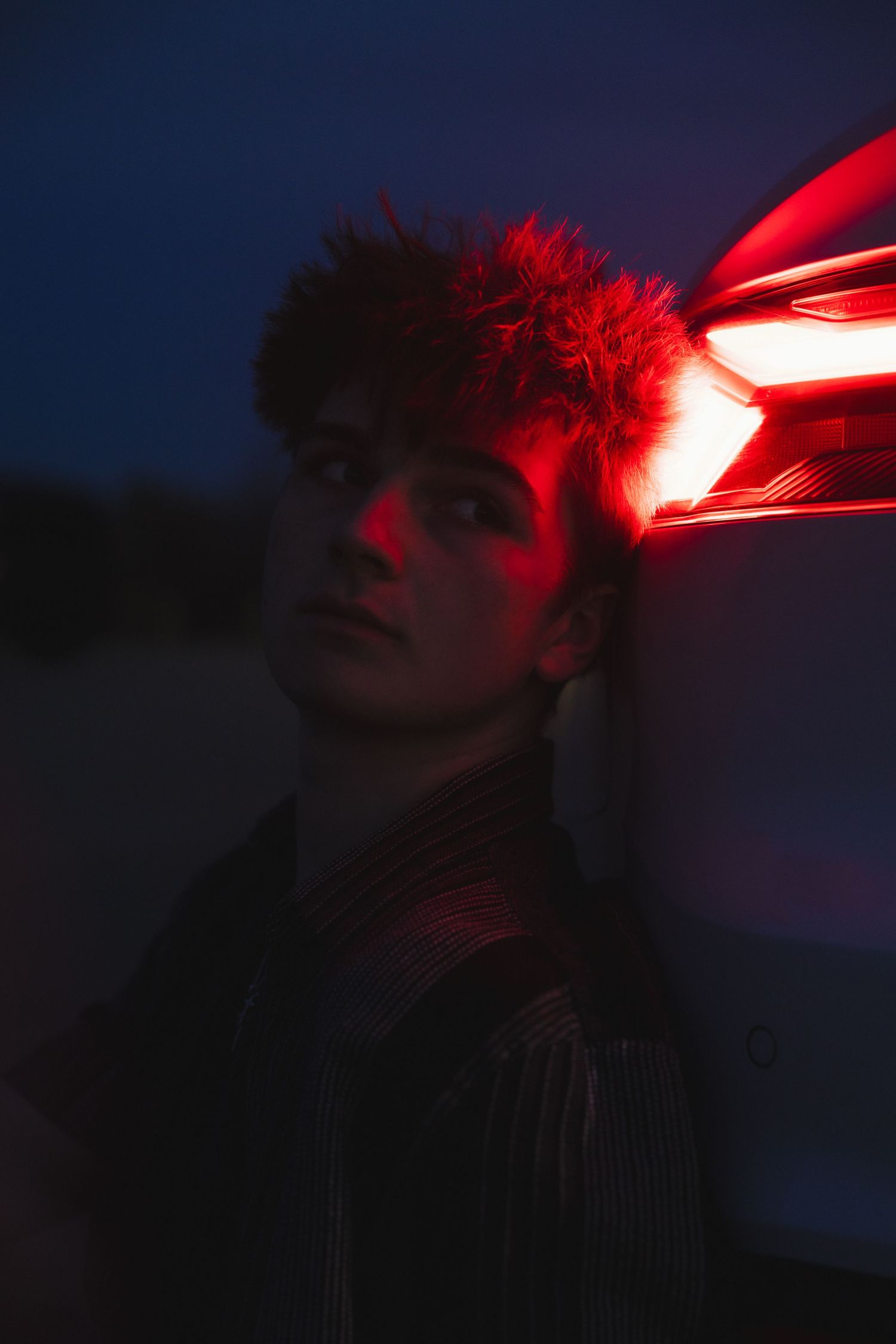 Young person leaning against a car taillight, bathed in dramatic red neon glow against dark background.