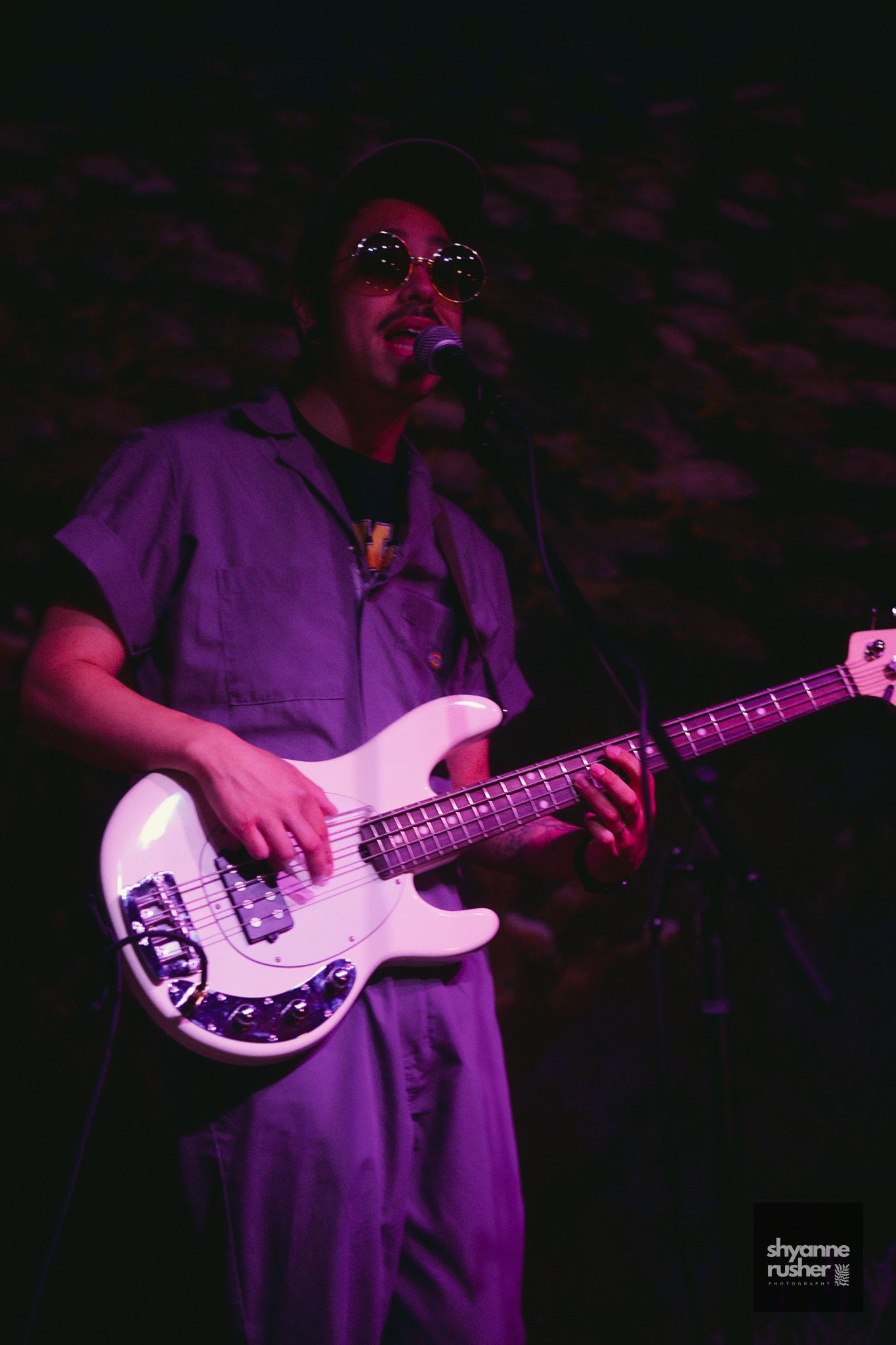 A series of darkly lit performance photos showing a musician playing a white bass guitar under purple stage lighting.