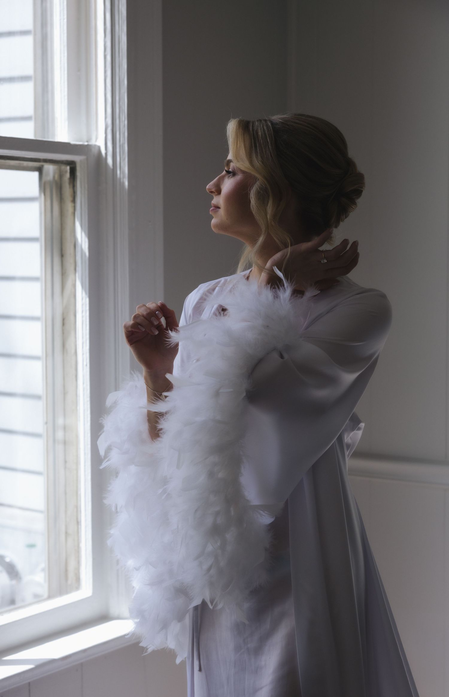 Bride in white feather robe gazing out a window in soft natural light on her wedding day.