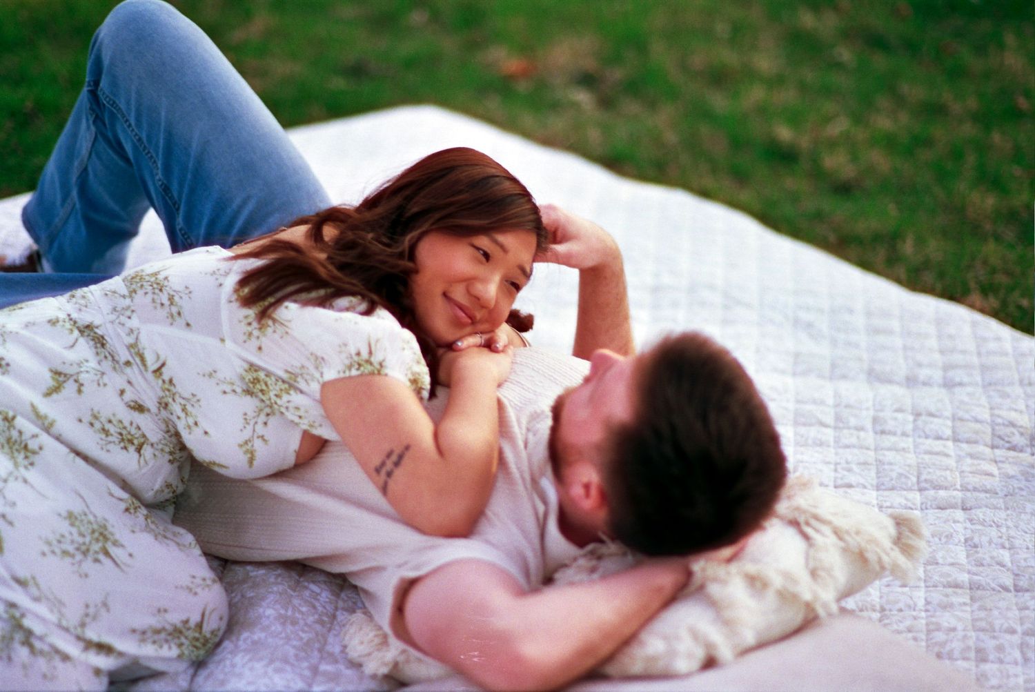 A couple lying together romantically on a white blanket in the grass.