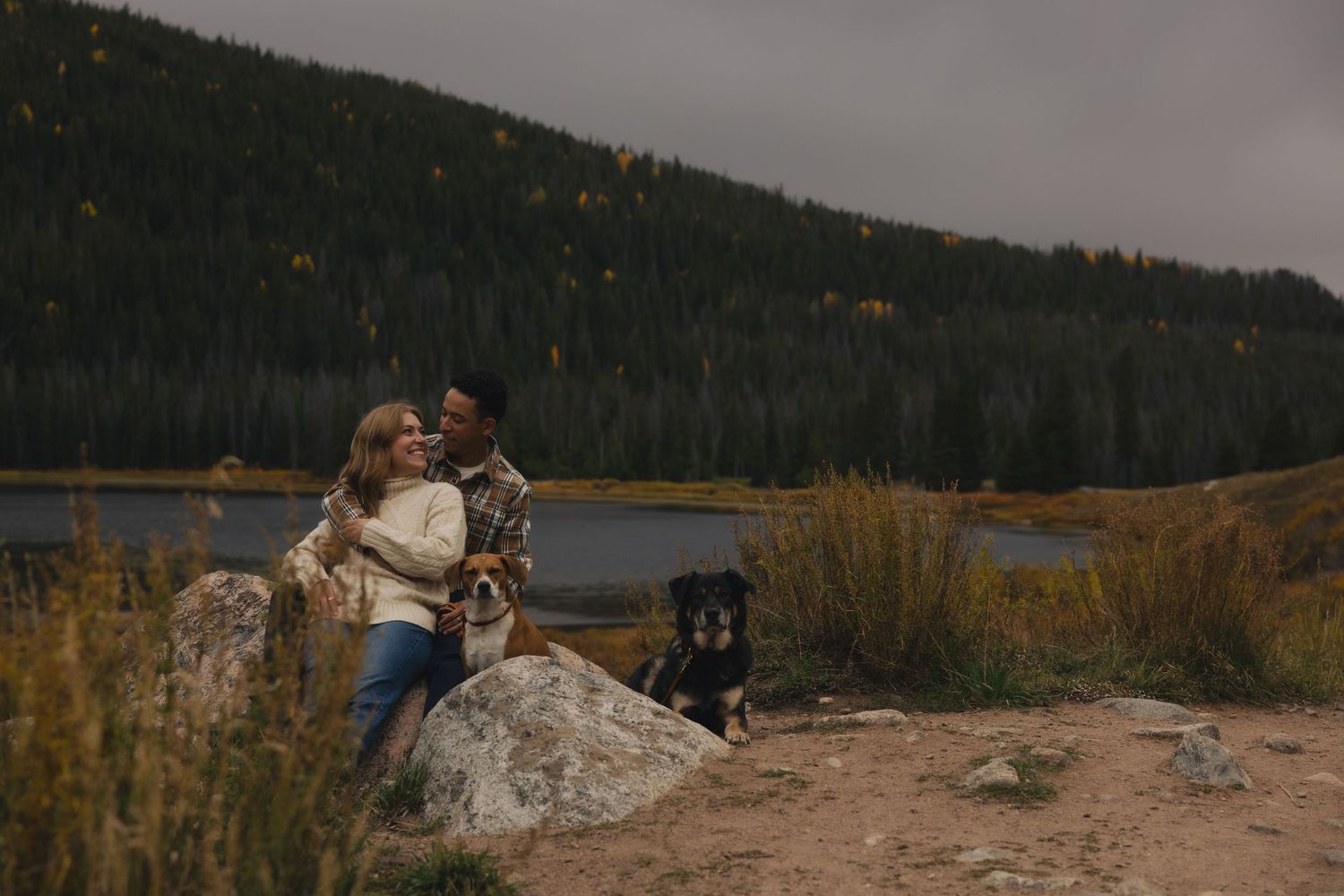 A couple shares a romantic moment on a scenic mountain overlook with a lake and forest in the background.