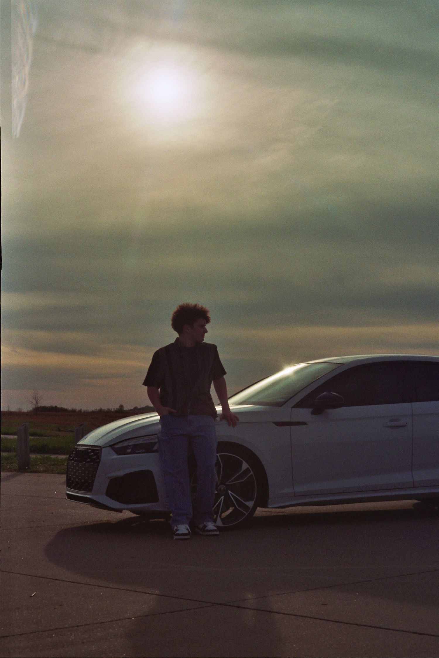 Young man in jeans and black tee leaning against a white Audi at sunset in an open parking lot.
