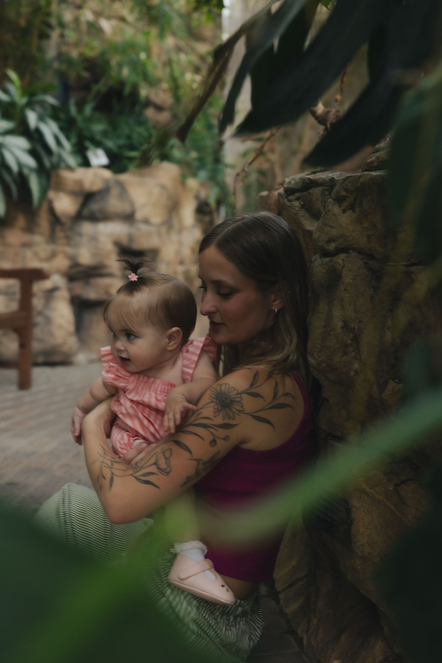 Tattooed mother tenderly holds her baby girl in pink dress among lush greenery and stone wall backdrop.