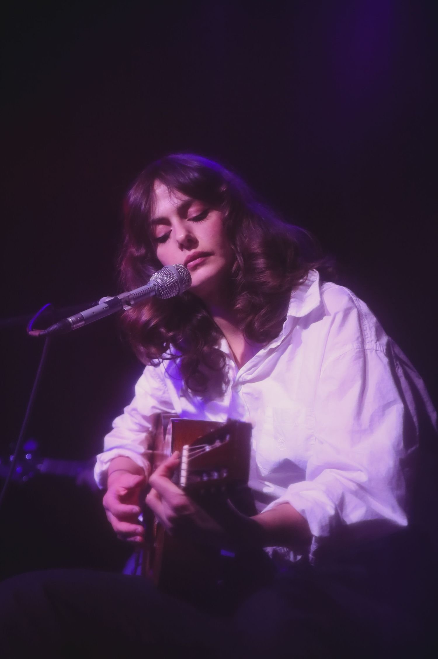 A performer in a white button-down shirt plays acoustic guitar and sings into a microphone on a dark stage with purple lighting.