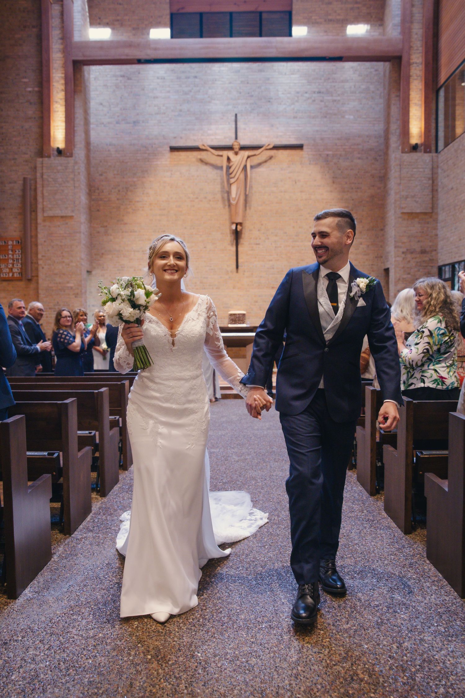 A newlywed couple walks down the aisle in a Catholic church with a large crucifix mounted on the brick wall behind them.