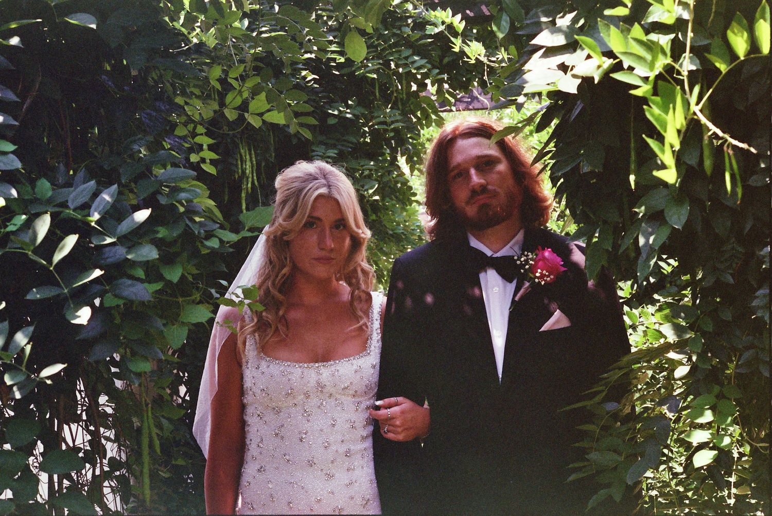 A couple in wedding attire poses together in a lush garden setting with greenery surrounding them.