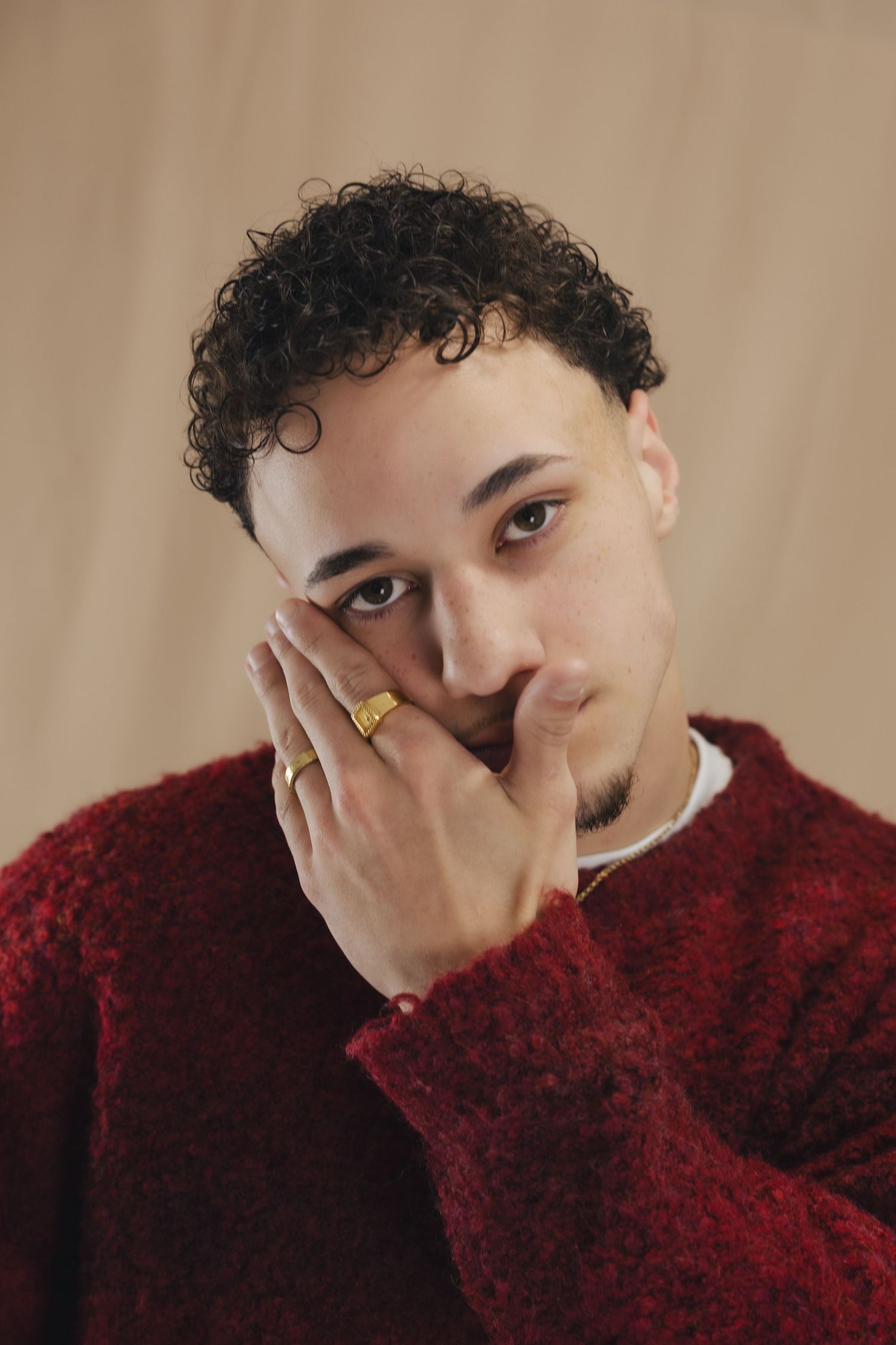 Young man with curly hair in red sweater, gold ring, hand on face, neutral background.