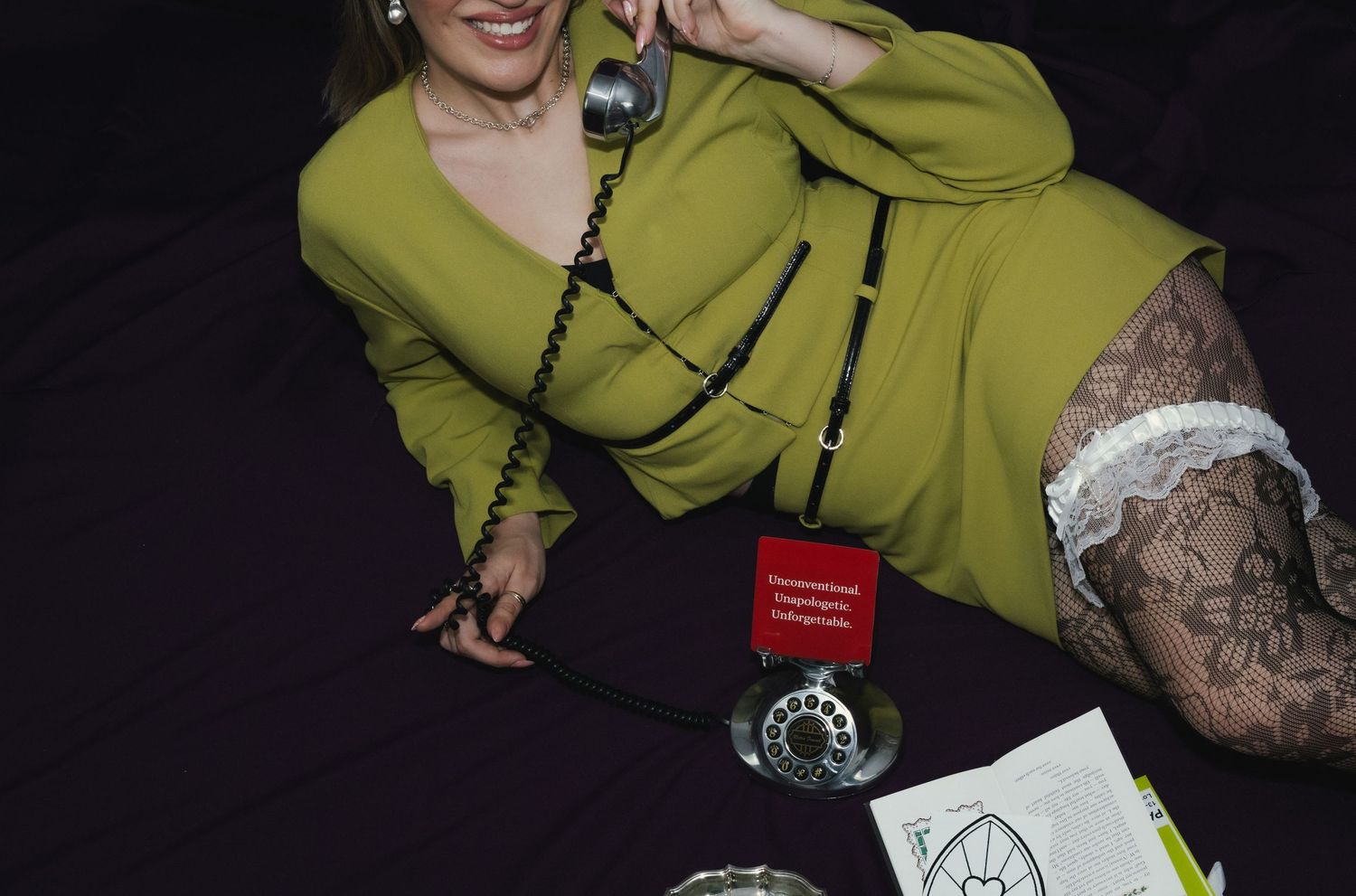 Woman in green suit lying down, holding vintage rotary phone, with tattoos and lace stockings visible.