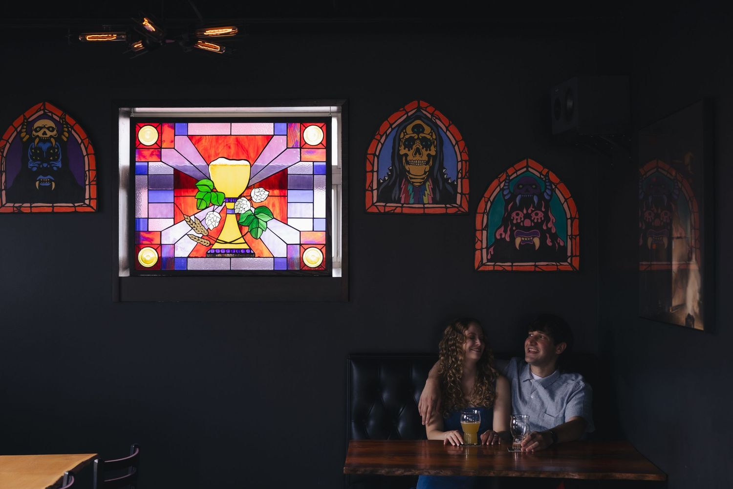 Couple sitting in a dark bar booth beneath colorful stained glass and skull artwork.