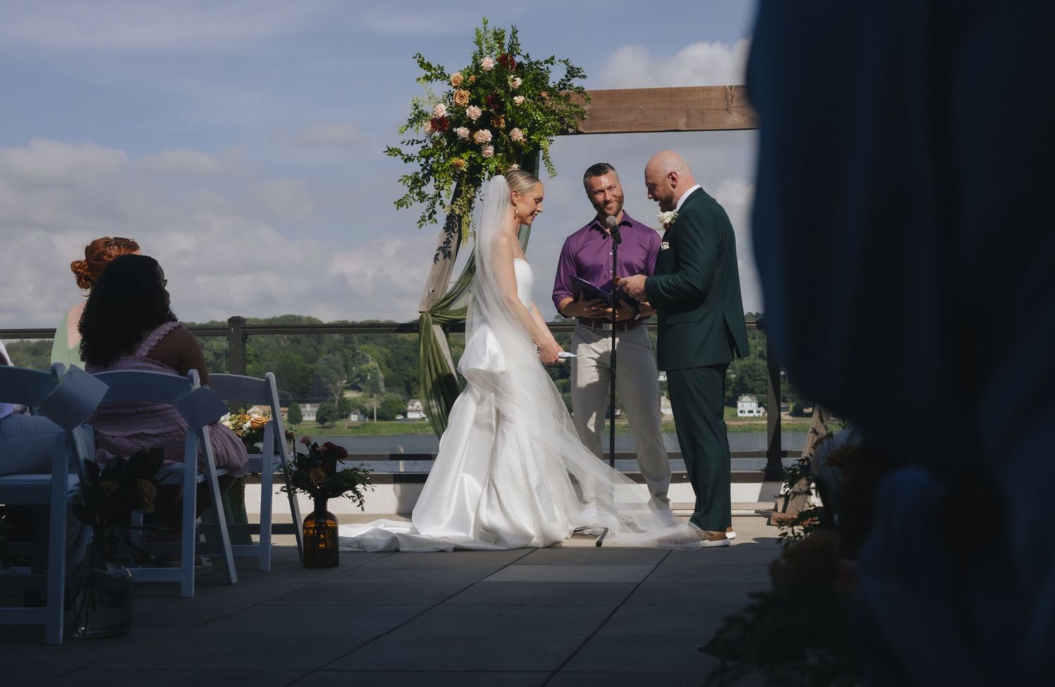 Outdoor rooftop wedding ceremony with bride in white gown and groom exchanging vows under floral arch.