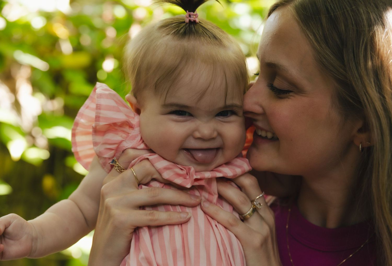 A joyful baby girl in a pink dress laughs while a smiling woman kisses her cheek outdoors.